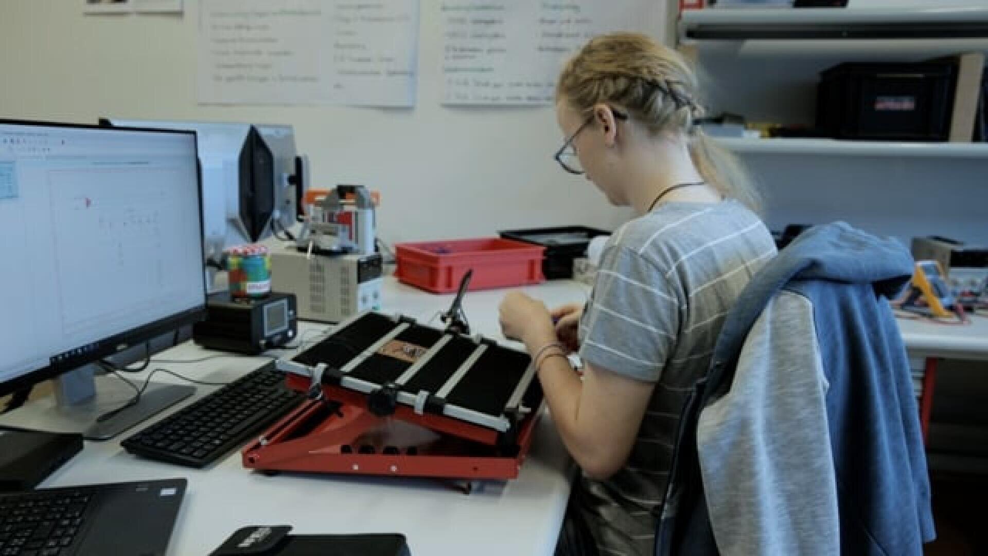 Eine Person mit blondem Haar in einem Zopf arbeitet an einem Schreibtisch mit elektronischen Bauteilen und einem Monitor, der ein technisches Diagramm anzeigt. Auf dem Schreibtisch befinden sich verschiedene Werkzeuge, eine Tastatur und geordnete Kisten.