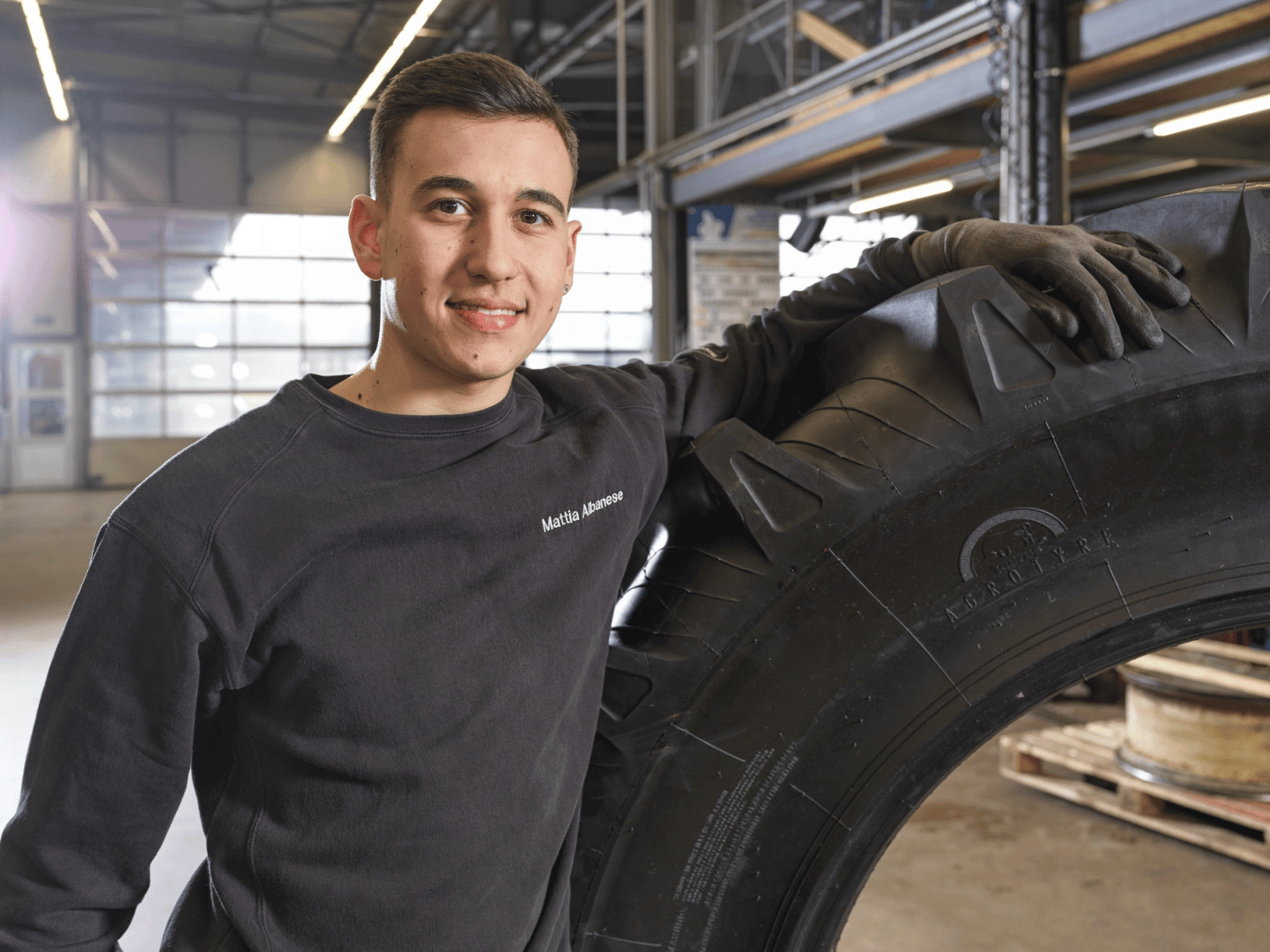 Ein junger Mann in Arbeitskleidung steht in einer geräumigen Werkstatt oder Garage, lächelt und stützt seinen behandschuhten Arm auf einen großen Industriereifen.