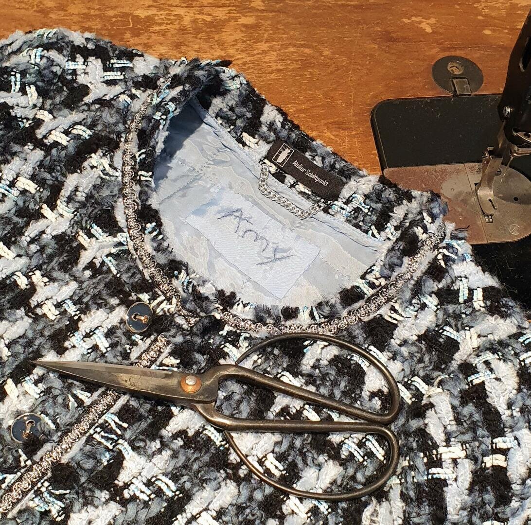 Eine Schere liegt auf einem strukturierten blauen, schwarzen und weißen Tweed-Kleidungsstück neben einer alten Nähmaschine auf einem Holztisch. Im Inneren des Kleidungsstücks ist ein Stoffetikett mit handgeschriebenem Text zu sehen.