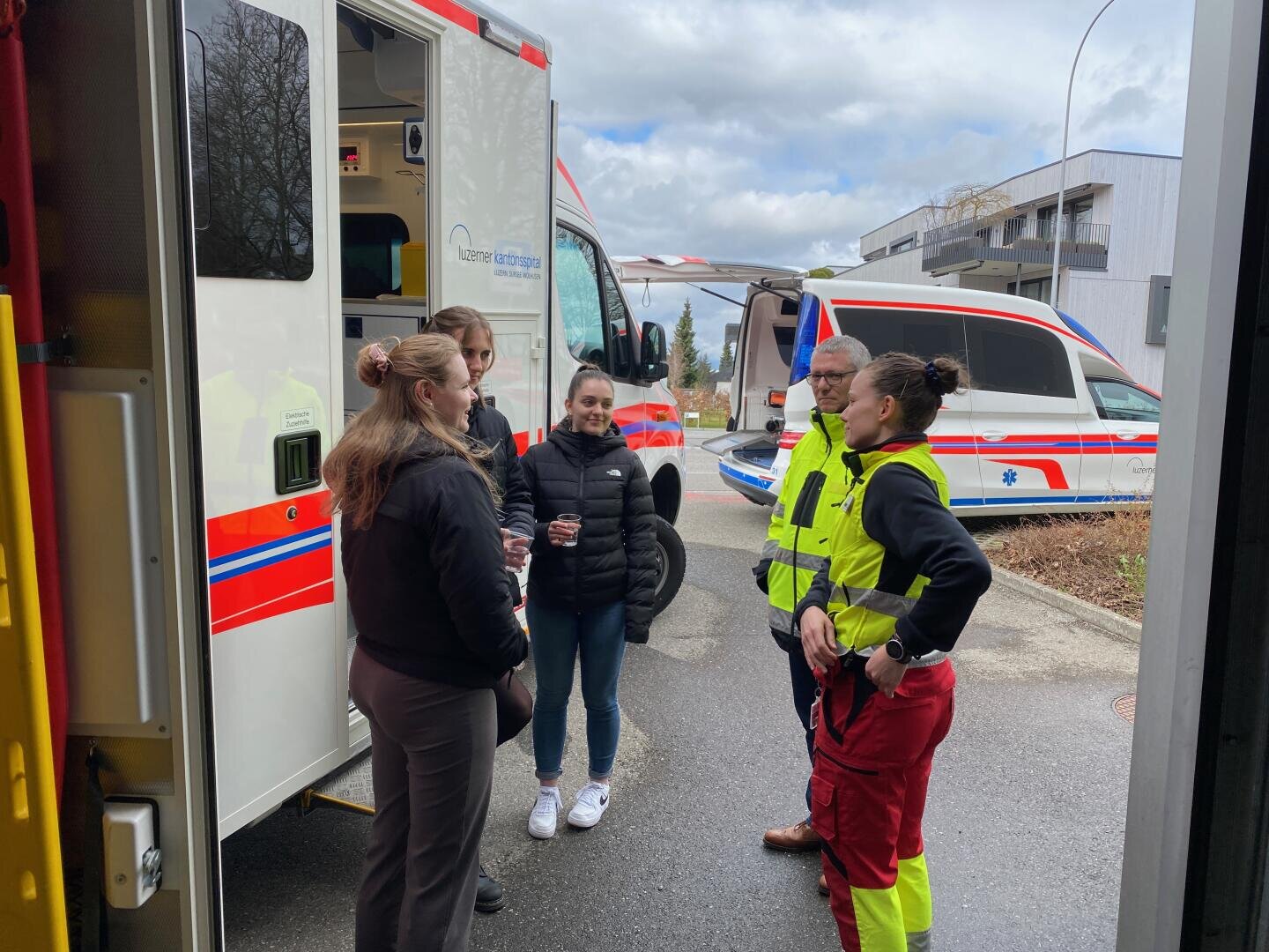 Zwei Sanitäter in gelben und roten Uniformen sprechen mit drei Frauen neben Krankenwagen, die an einem bewölkten Tag draußen geparkt sind. Die Gruppe steht neben der offenen Tür eines Rettungswagens.