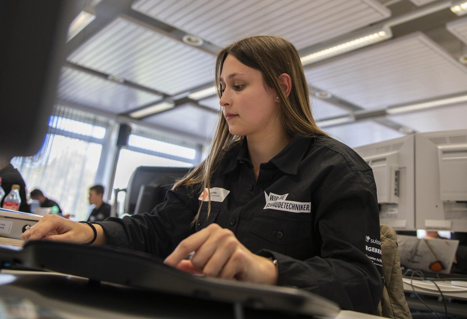 Eine junge Frau in einem schwarzen Shirt arbeitet an einem Computer in einem hellen, modernen Büro, im Hintergrund sind weitere Personen und Geräte zu sehen.