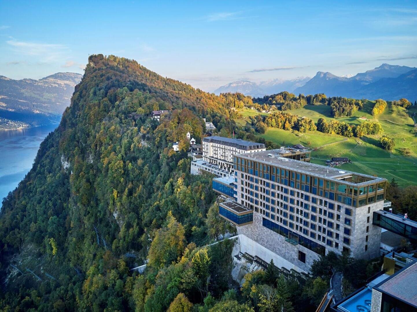 Ein modernes Hotel mit großen Fenstern liegt an einem bewaldeten Berghang mit Blick auf einen See, mit sanften grünen Hügeln und fernen Berggipfeln unter einem klaren blauen Himmel.