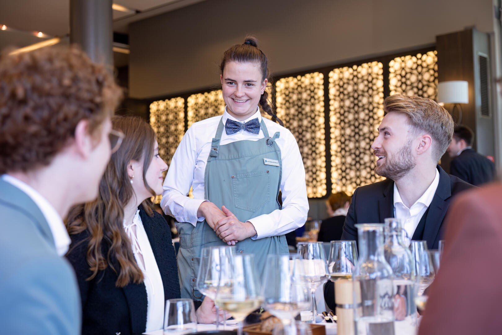 Eine lächelnde Kellnerin mit Fliege und Schürze steht an einem Tisch und unterhält sich mit vier sitzenden Gästen in einem modernen Restaurant mit dekorativer Verkleidung und Weingläsern auf dem Tisch.