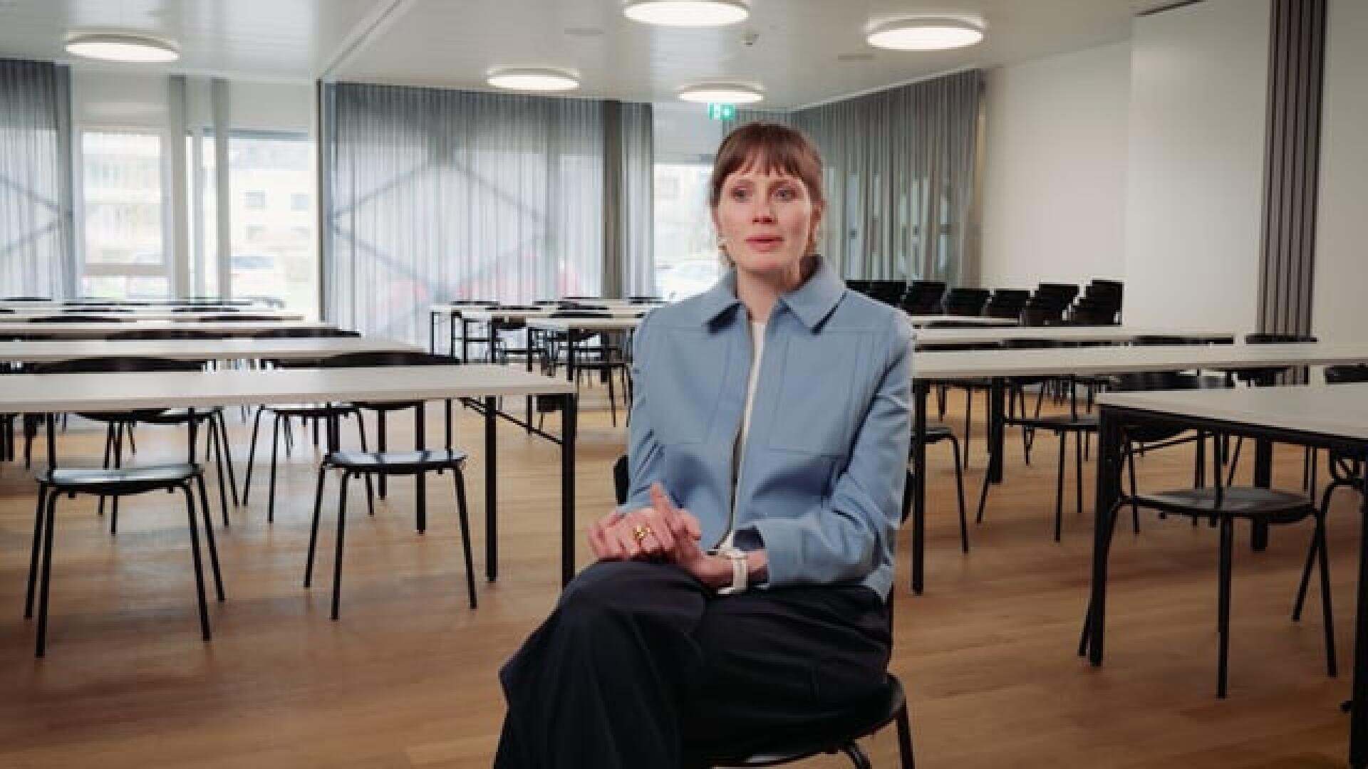 Eine Frau in einer hellblauen Jacke sitzt auf einem Stuhl in einem hellen, leeren Klassenzimmer, hinter ihr stehen Tische und Stühle ordentlich aufgereiht.