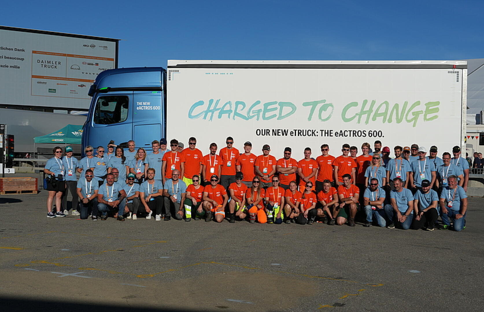 Eine große Gruppe von Menschen in passenden blauen und orangefarbenen Hemden posiert gemeinsam vor einem blau-weißen LKW mit dem Slogan Charged to Change und Our New eTruck: Der eActros 600 auf seiner Seite.