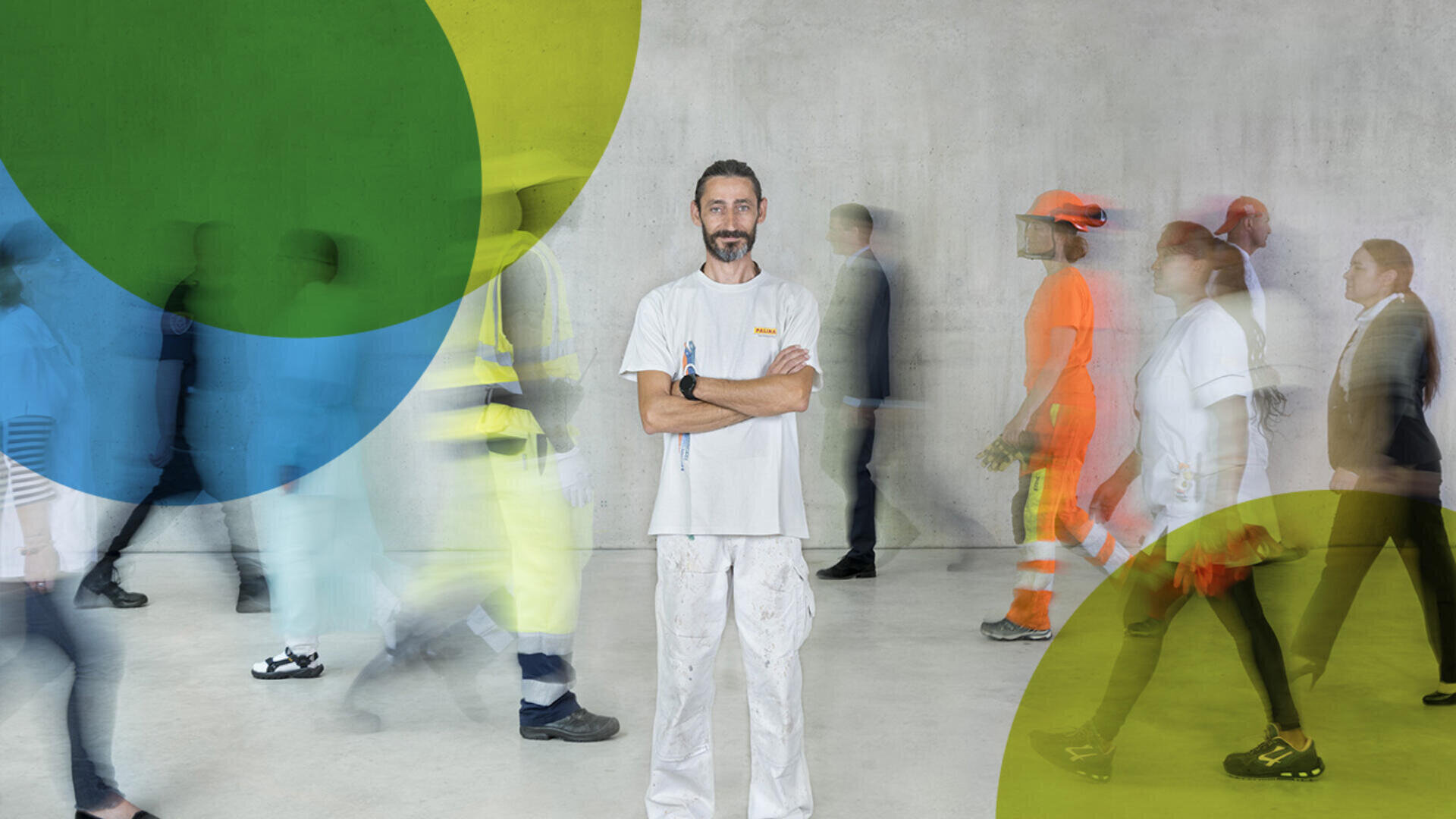 Ein Mann in weißer Arbeitskleidung steht selbstbewusst mit verschränkten Armen da, während verschwommene Gestalten in verschiedenen Uniformen und Geschäftskleidung an ihm vorbei gegen eine graue Wand mit großen transparenten grünen und blauen Kreisen laufen.