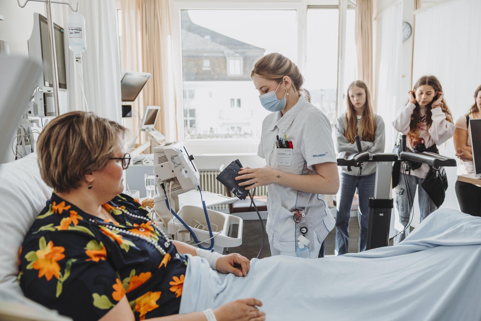 Eine Krankenschwester mit Maske prüft den Blutdruck eines Patienten in einem Krankenhauszimmer, während drei Besucher in der Nähe stehen und die Szene beobachten. Der Patient liegt im Bett, trägt eine Brille und ein geblümtes Hemd.