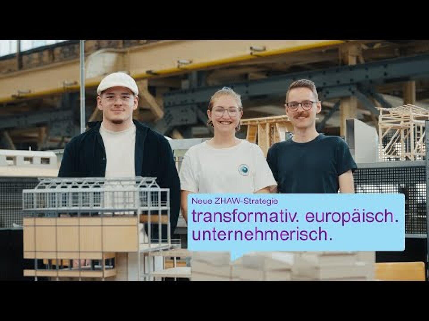 Drei junge Erwachsene stehen lächelnd hinter Architekturmodellen in einem Innenraum. In einem blauen Textkasten steht in deutscher Sprache: Neue ZHAW-Strategie: transformativ. europäisch. unternehmerisch. Industrieller Hintergrund sichtbar.