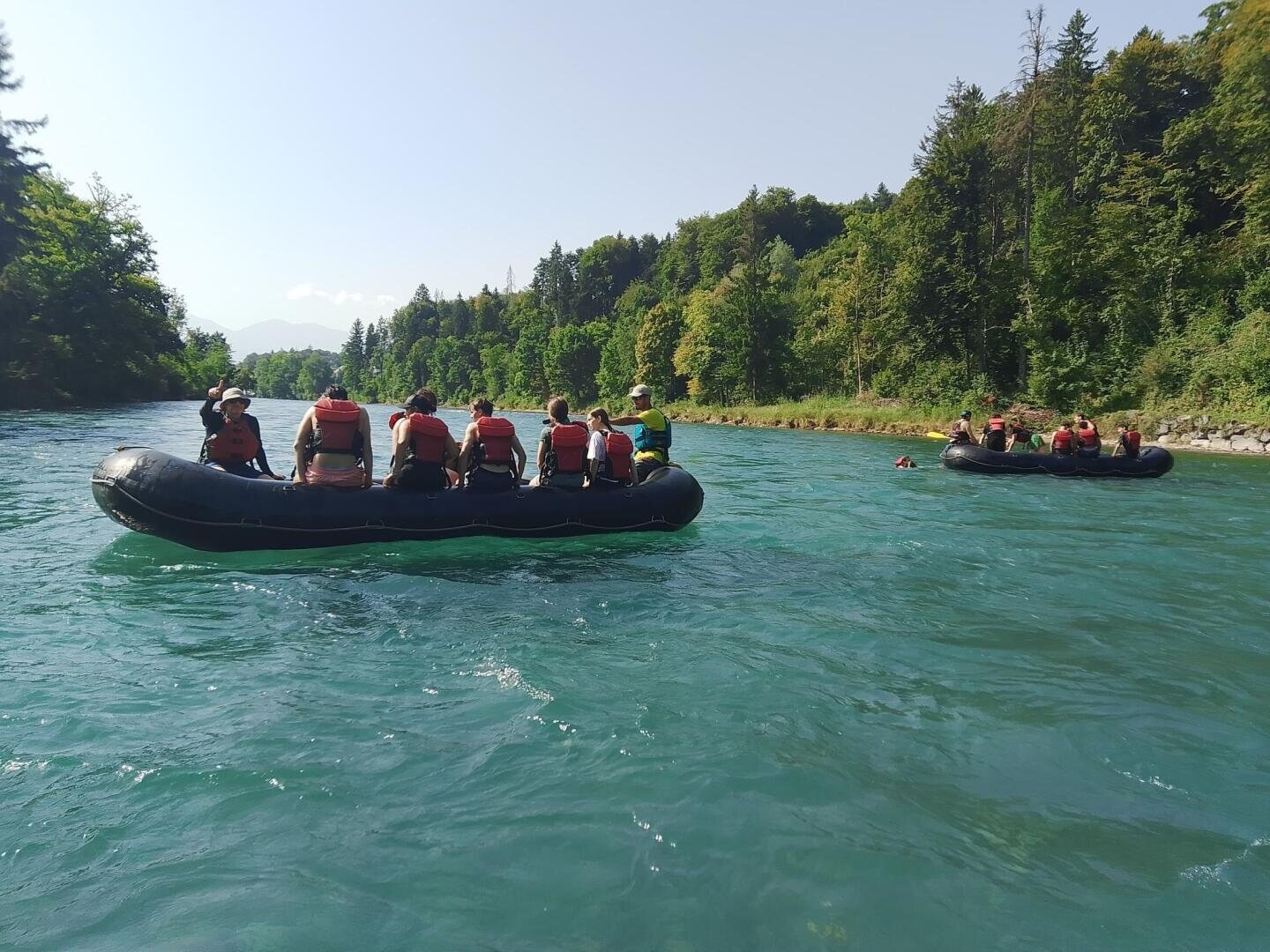 Zwei aufblasbare Flöße mit Personen, die Schwimmwesten tragen, treiben auf einem blauen Fluss, umgeben von üppig grünen Bäumen und Hügeln, unter einem klaren Himmel.