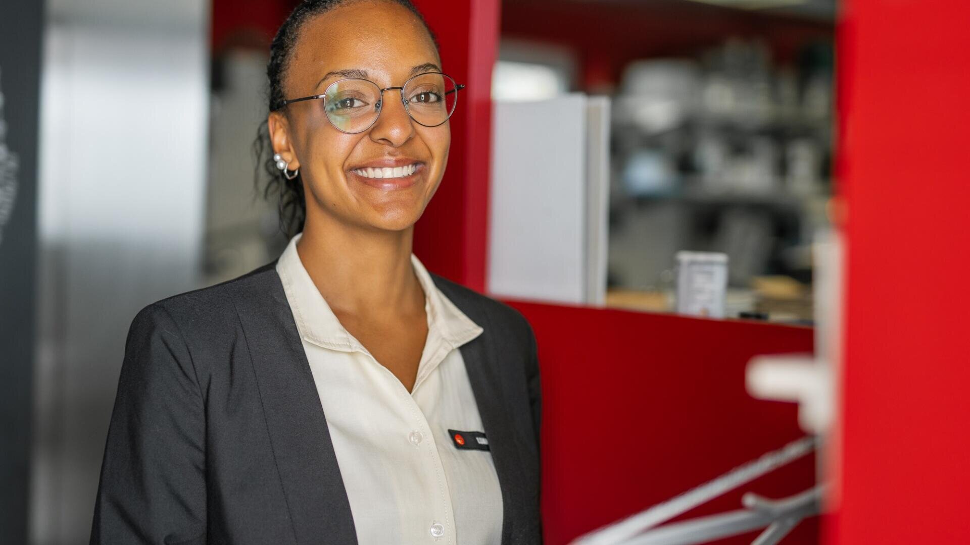 Eine Frau, die eine Brille, einen schwarzen Blazer und eine helle Bluse trägt, lächelt, während sie in einem modernen Büro mit roten und weißen Wänden im Hintergrund steht.