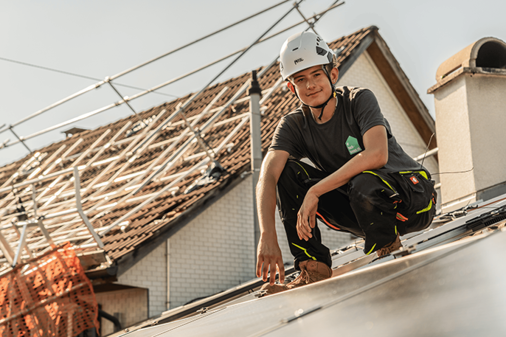 Eine Person mit Helm und Arbeitskleidung hockt auf einem im Bau befindlichen Dach, im Hintergrund sind Gerüste und Dachmaterialien zu sehen.