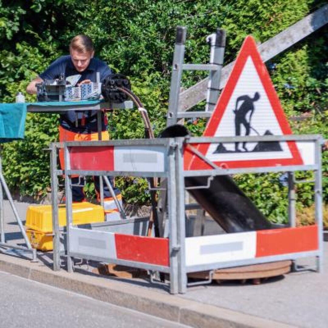 Ein Arbeiter steht in der Nähe eines umzäunten Baustellenbereichs mit einem Bauschild, Werkzeugen und Geräten auf einem Tisch, umgeben von Grünpflanzen.