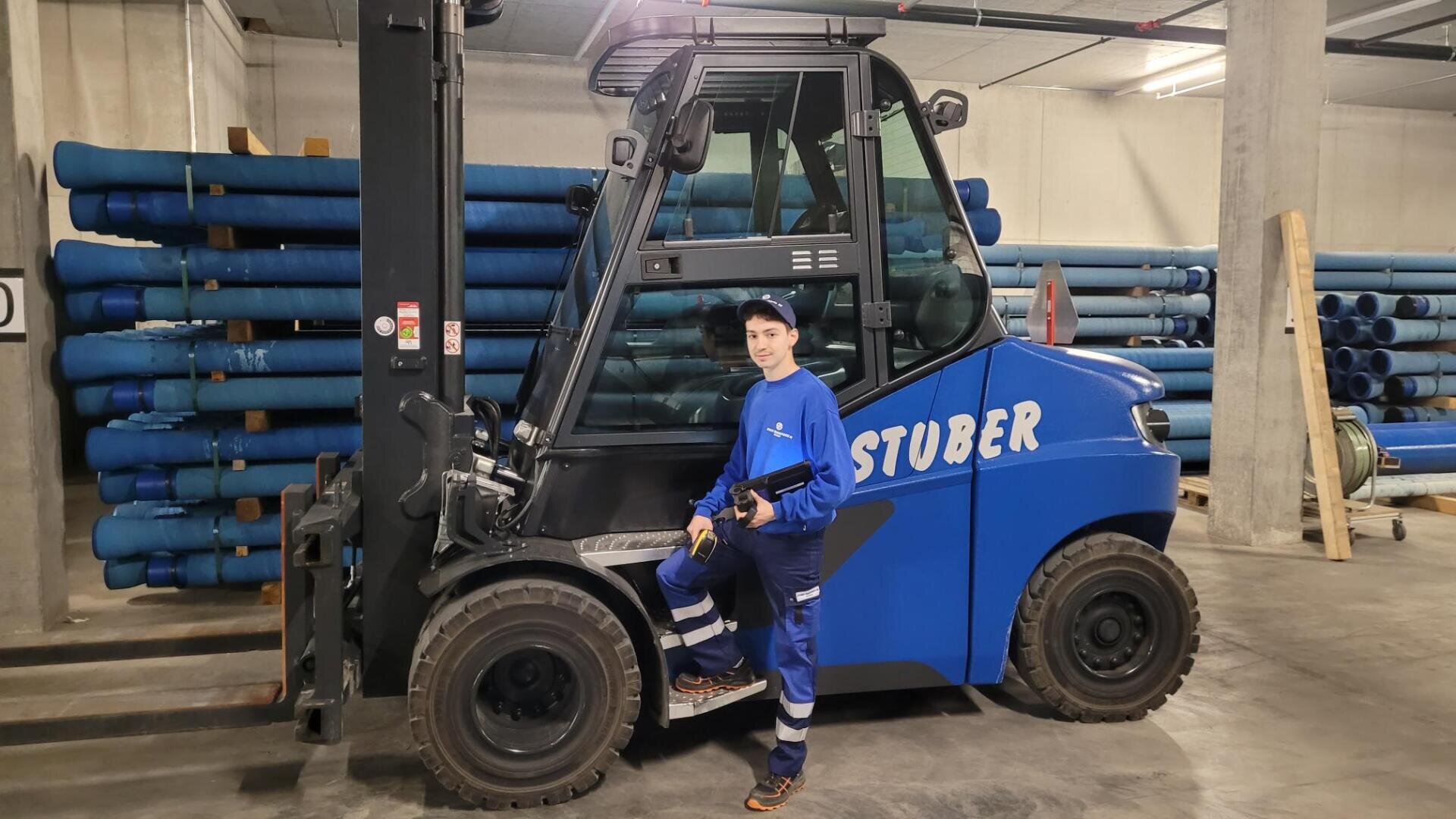 Eine Person in blauer Arbeitskleidung und mit Mütze steht neben einem großen blauen Gabelstapler mit der Aufschrift STUBER in einem Lagerhaus mit gestapelten blauen Rohren im Hintergrund.