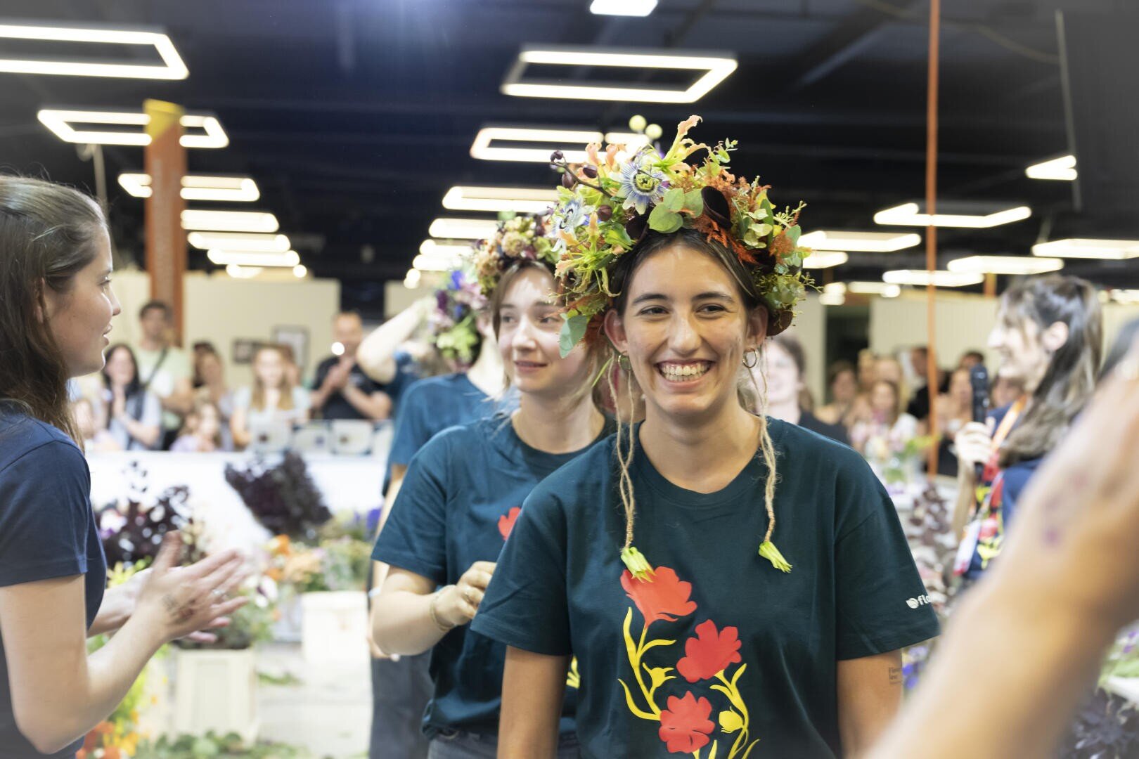 Eine junge Frau trägt eine Blumenkrone und lächelt strahlend, während sie in einem belebten Innenraum mit anderen um sie herum steht. Sie und andere tragen dunkle Hemden mit bunten Blumenmustern.