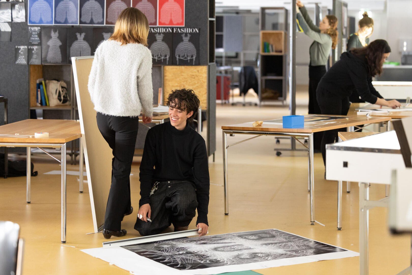 Ein junger Mann kniet auf dem Boden und lächelt, während er an einem großen Schwarz-Weiß-Kunstwerk arbeitet. Eine Frau steht neben ihm, und im Hintergrund arbeiten mehrere Personen in einem geräumigen, kreativen Atelier.