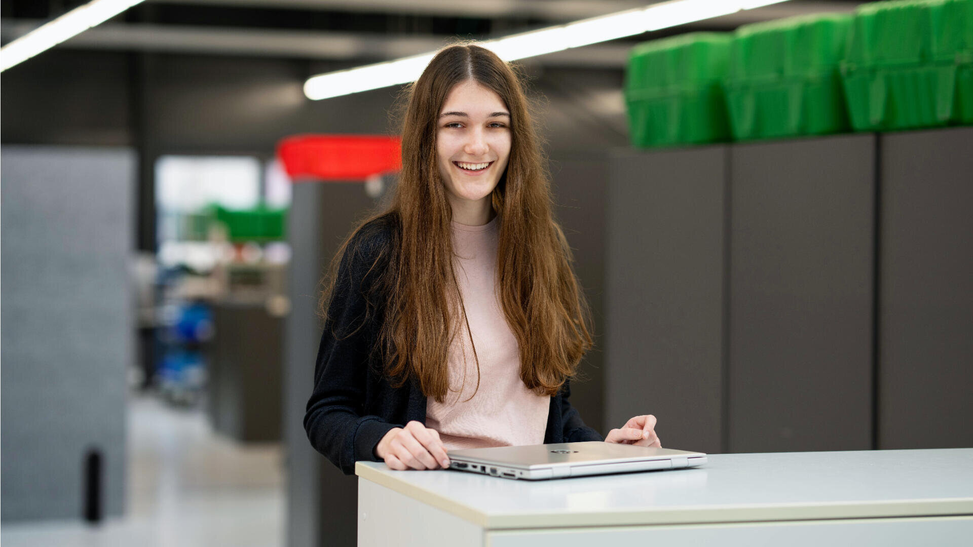 Eine junge Frau mit langen braunen Haaren, die ein hellrosa Oberteil und eine dunkle Strickjacke trägt, lächelt, während sie an einem weißen Schreibtisch mit geschlossenem Laptop in einer modernen Büro- oder Arbeitsumgebung steht.