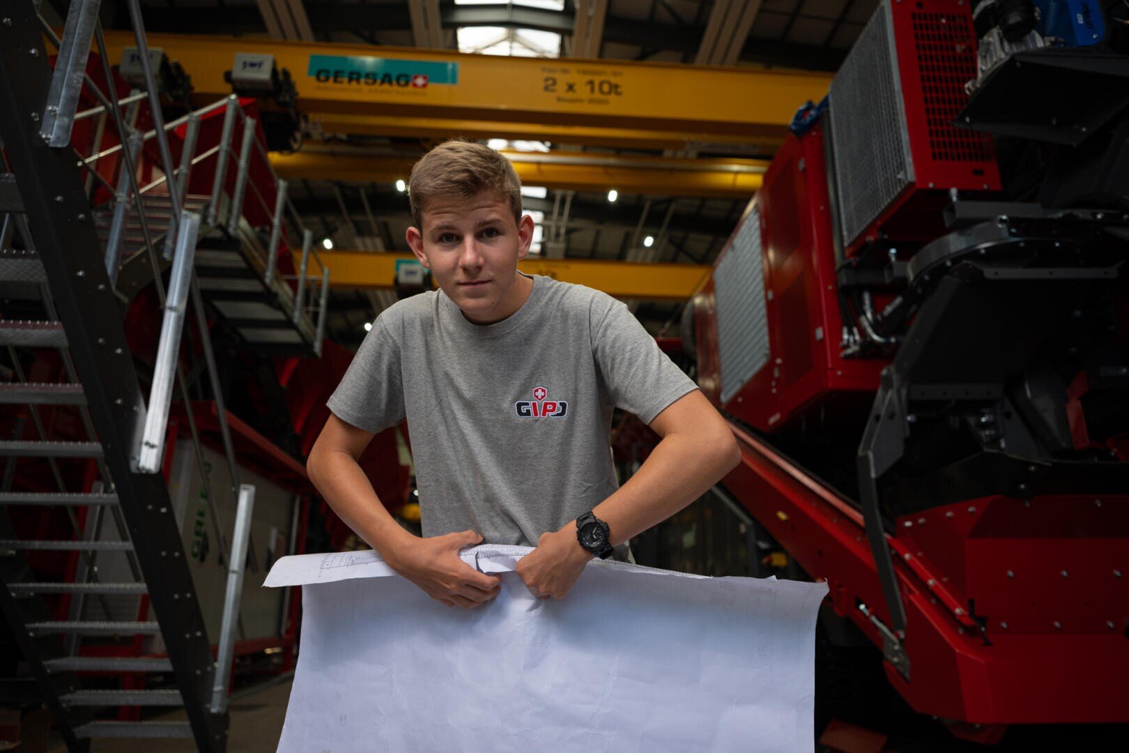 Ein junger Mann in einem grauen GIPO-Shirt hält ein großes Blatt Papier in einer Industriehalle in der Hand, im Hintergrund sind rote Maschinen und ein gelber Laufkran zu sehen.