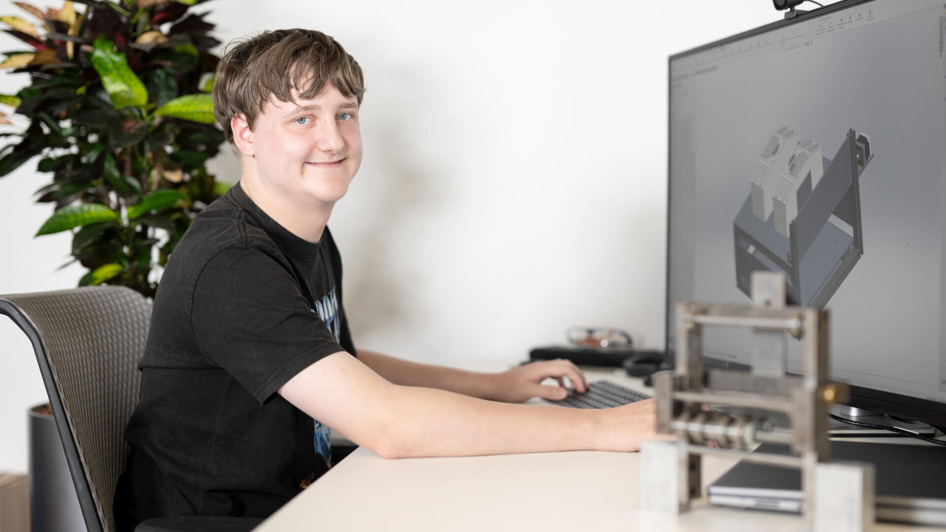 Ein junger Mensch sitzt an einem Schreibtisch und lächelt in die Kamera, während er einen Computer mit einem 3D-Modellierungsprogramm auf dem Bildschirm benutzt. Im Arbeitsbereich sind ein Metallobjekt und eine Topfpflanze zu sehen.