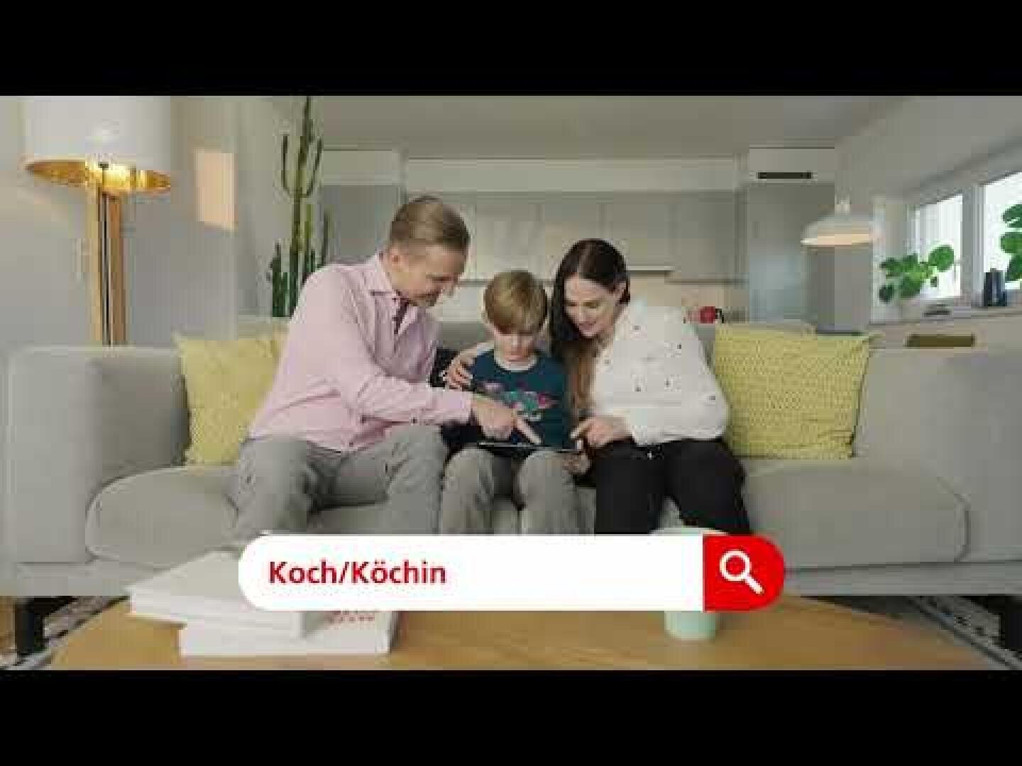 Eine lächelnde dreiköpfige Familie sitzt auf einer grauen Couch in einem modernen Wohnzimmer und schaut gemeinsam auf ein Tablet. Auf dem Bildschirm ist eine rote Suchleiste mit dem Text Koch/Köchin und einem Lupensymbol zu sehen.