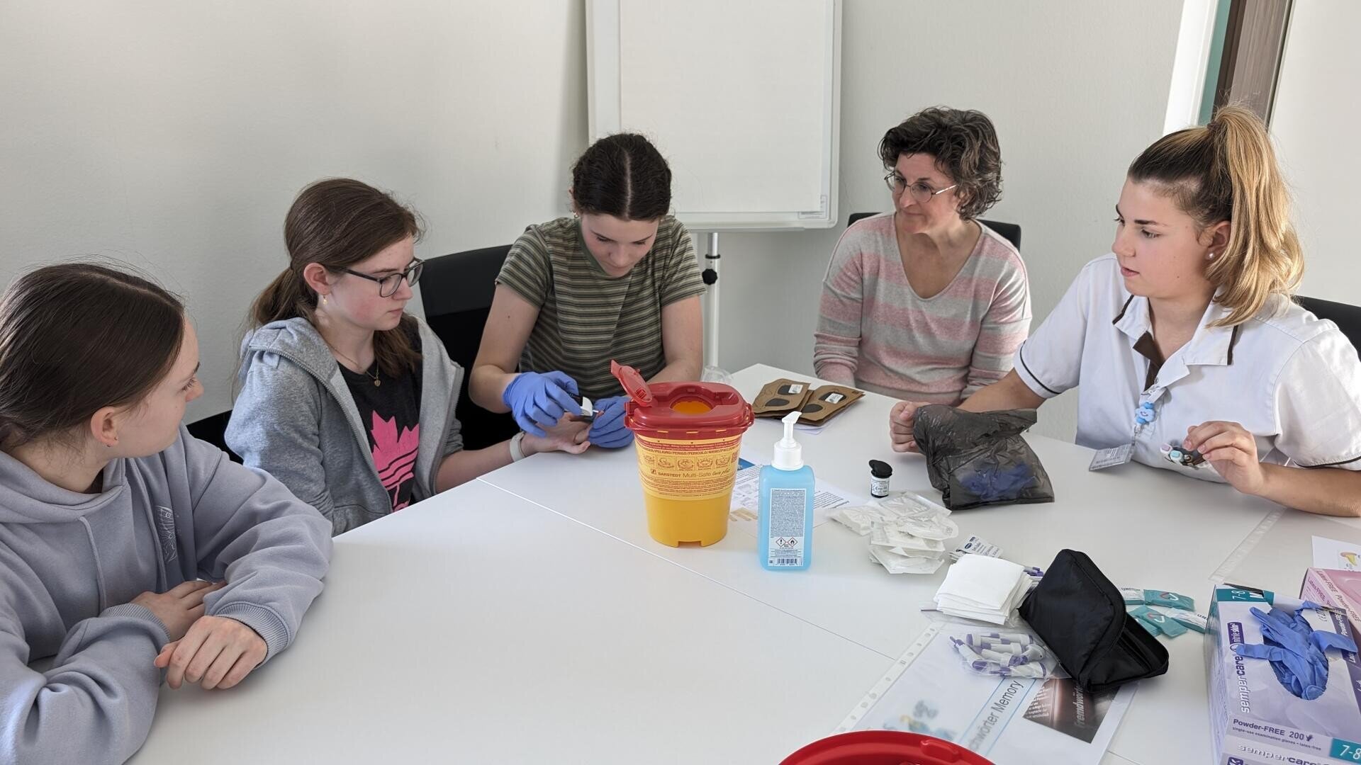 Fünf Frauen sitzen um einen Tisch mit medizinischem Material. Eine Frau mit Handschuhen demonstriert einen Eingriff an einem anderen Finger, während die anderen aufmerksam zusehen. Rechts sitzt eine Krankenschwester in Weiß, und auf dem Tisch steht ein gelber Behälter für scharfe Gegenstände.