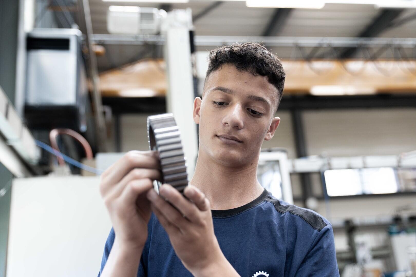 Ein junger Mann in einer Werkstatt untersucht ein großes Metallgetriebe, das er mit beiden Händen hält, und trägt ein blaues T-Shirt. Im Hintergrund sind Industrieanlagen und Werkzeuge zu sehen.