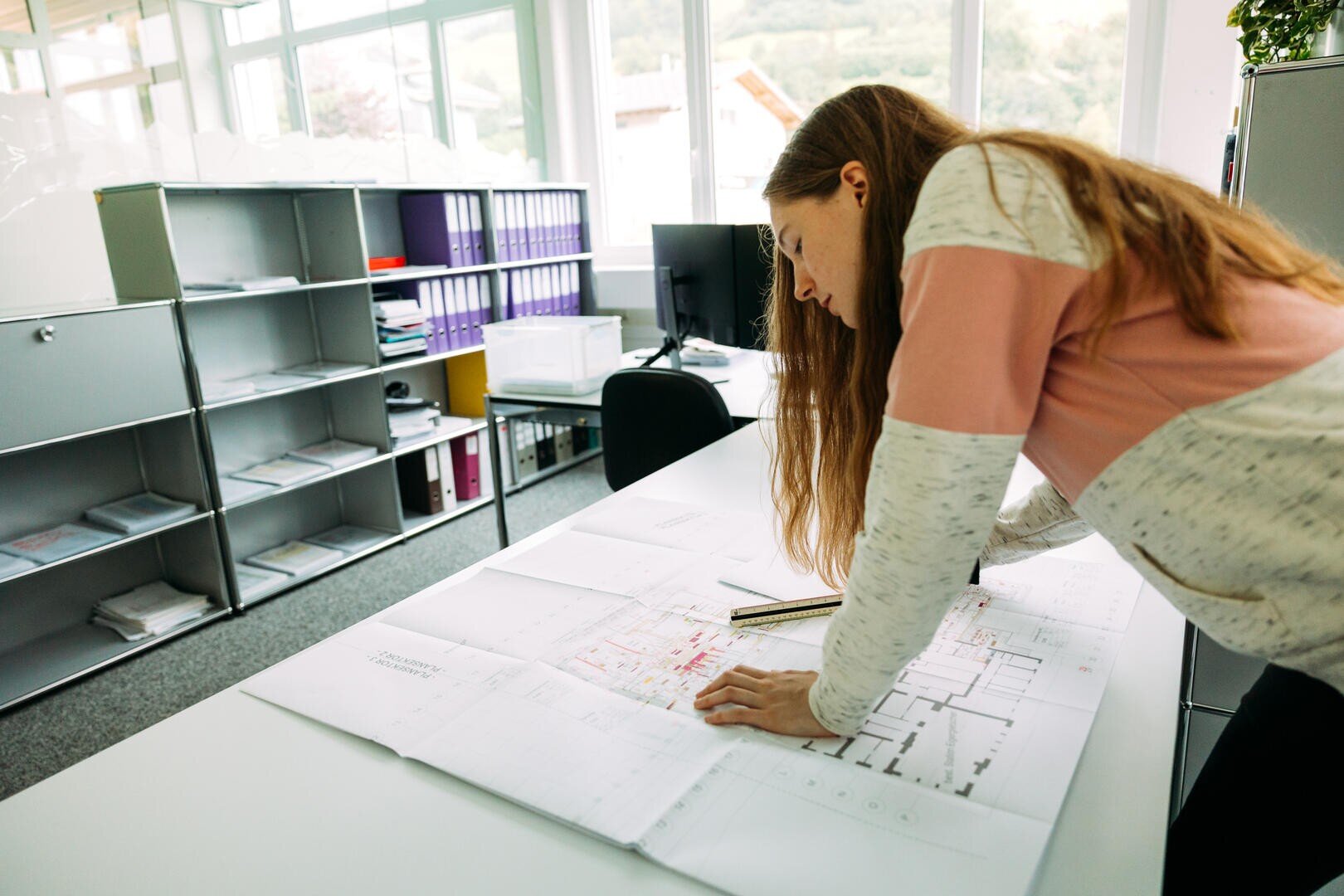 Eine Frau mit langen Haaren beugt sich über einen Schreibtisch und prüft in einem hellen Büro mit Regalen, Ordnern und großen Fenstern im Hintergrund Baupläne.