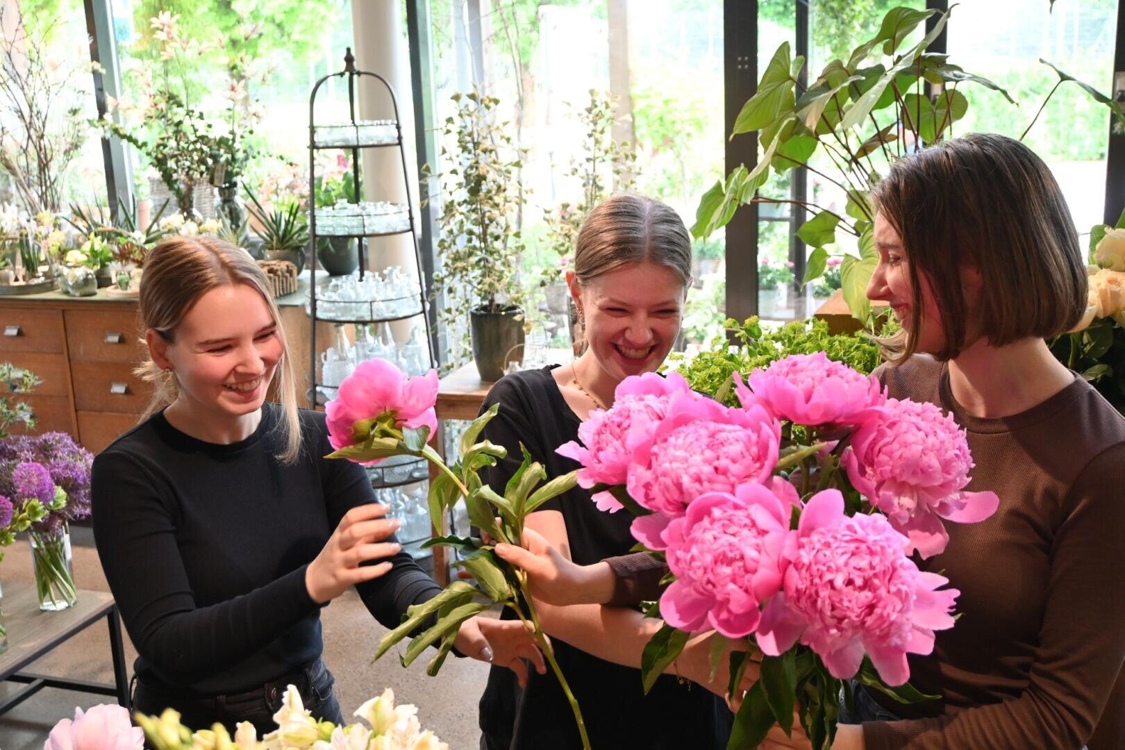 Drei Frauen in einem Blumenladen lächeln und lachen, während sie große rosa Pfingstrosen in den Händen halten, umgeben von grünen Pflanzen und Blumenarrangements.