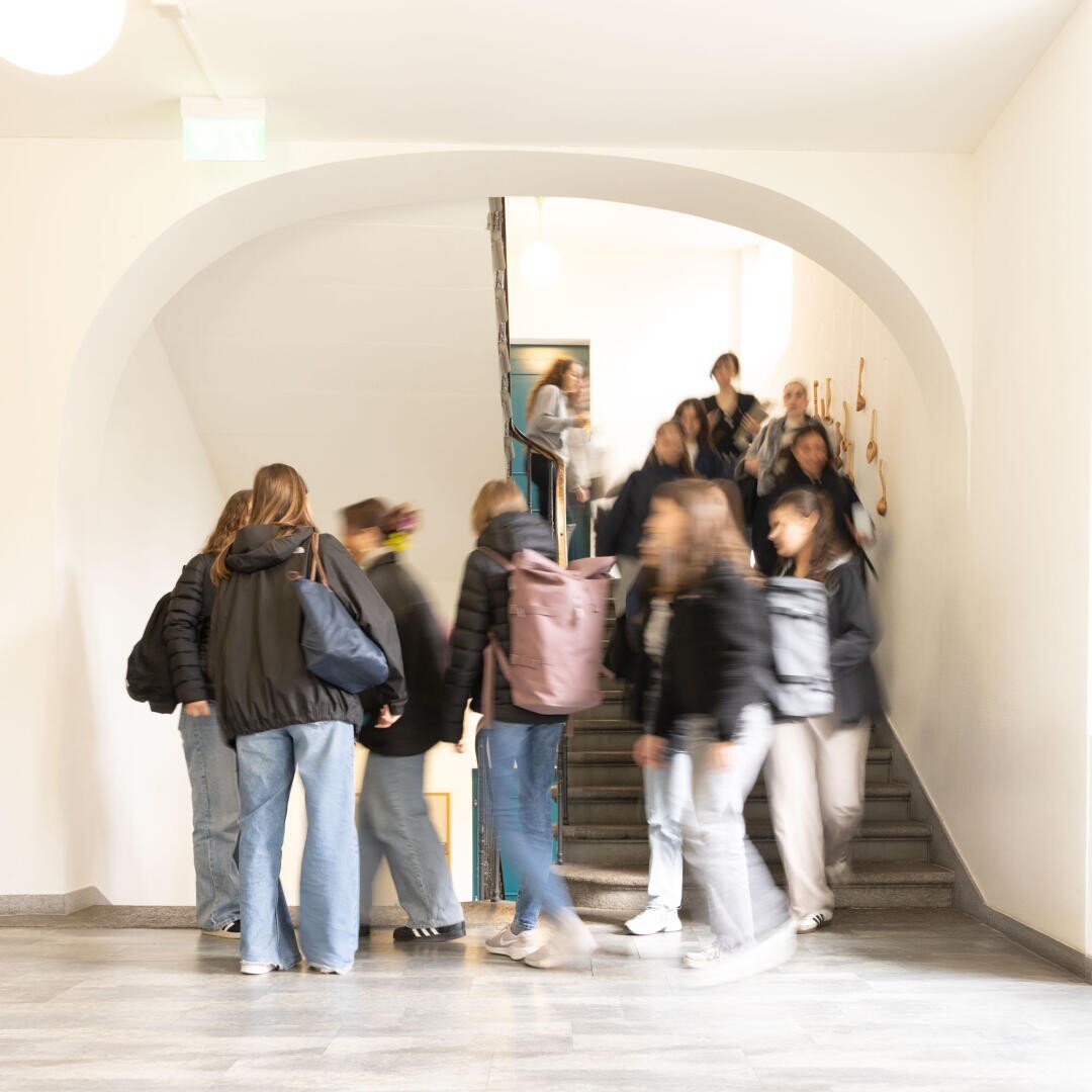 Eine Gruppe von Schülern in Freizeitkleidung geht in einem hellen Schulflur eine Treppe hinauf und hinunter, wobei ihre Bewegungen leicht unscharf sind, um Bewegung zu zeigen.