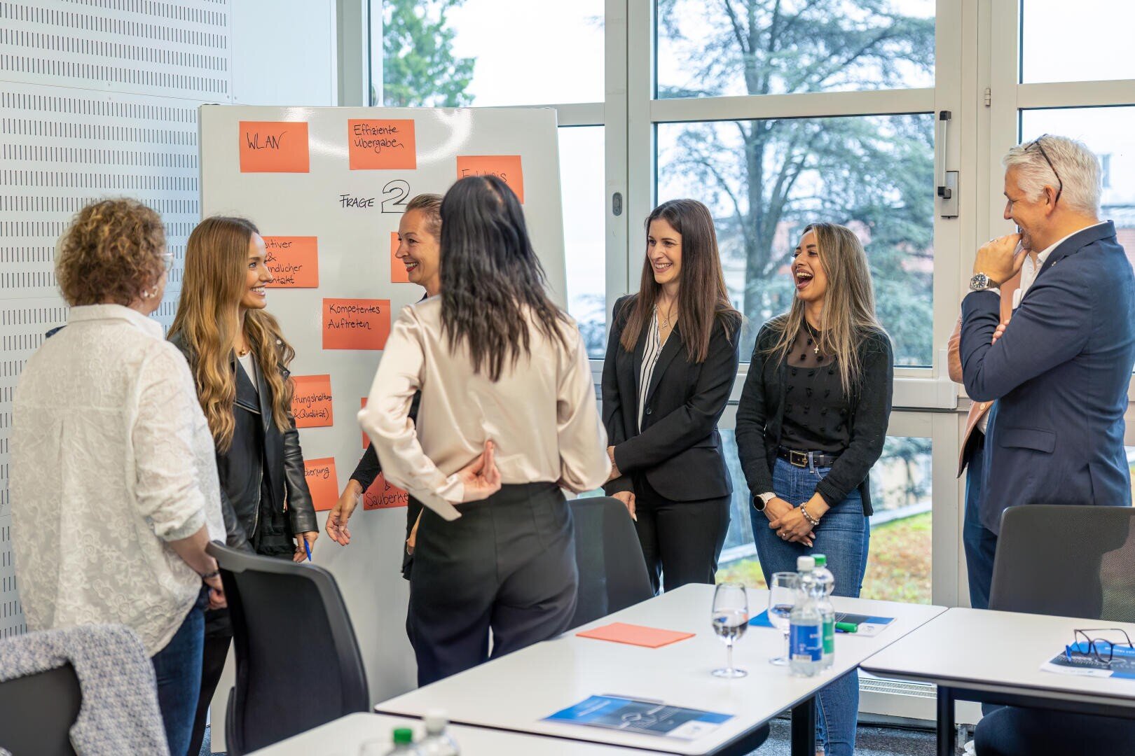 Sechs Personen, vier Frauen und zwei Männer, stehen in einem hellen Büroraum um ein Whiteboard mit orangefarbenen Haftnotizen herum, lächeln und diskutieren. In der Nähe stehen Tische mit Papieren und Wasserflaschen.