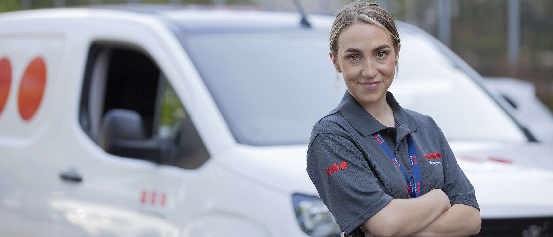 Ein Lieferfahrer steht selbstbewusst mit verschränkten Armen vor einem weißen Firmenwagen und trägt eine graue Uniform sowie einen Ausweis.