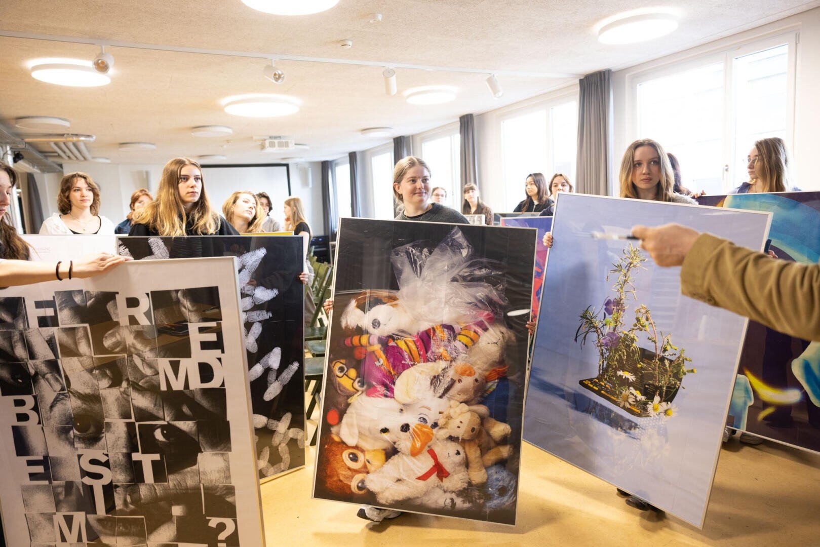 Eine Gruppe junger Menschen hält in einem hellen Raum große Kunstwerke hoch, darunter eine Collage aus Buchstaben, einen Stapel ausgestopfter Tiere und ein Blumenarrangement. Eine Person im Vordergrund gestikuliert in Richtung der ausgestellten Kunstwerke.