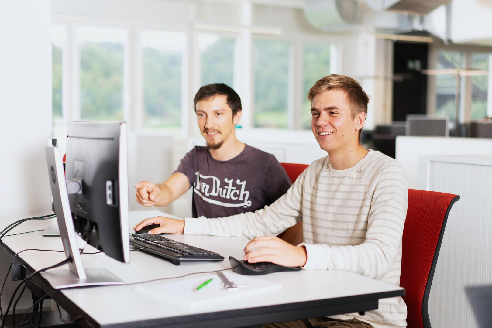 Zwei Männer sitzen an einem Schreibtisch und schauen auf einen Computerbildschirm, während einer von ihnen auf den Monitor zeigt.