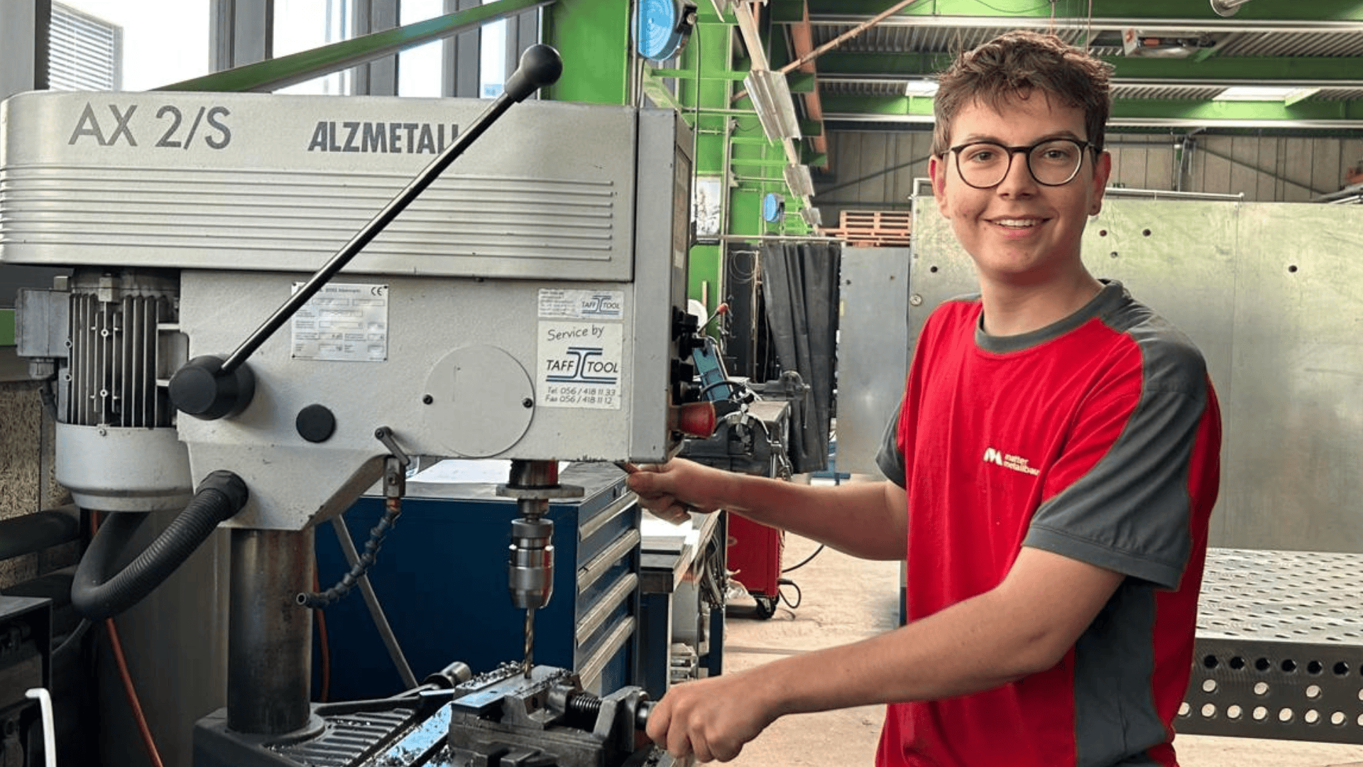 Ein junger Mann mit Brille und rotem Hemd bedient in einer Werkstatt eine Bohrmaschine der Marke Alzmetall.