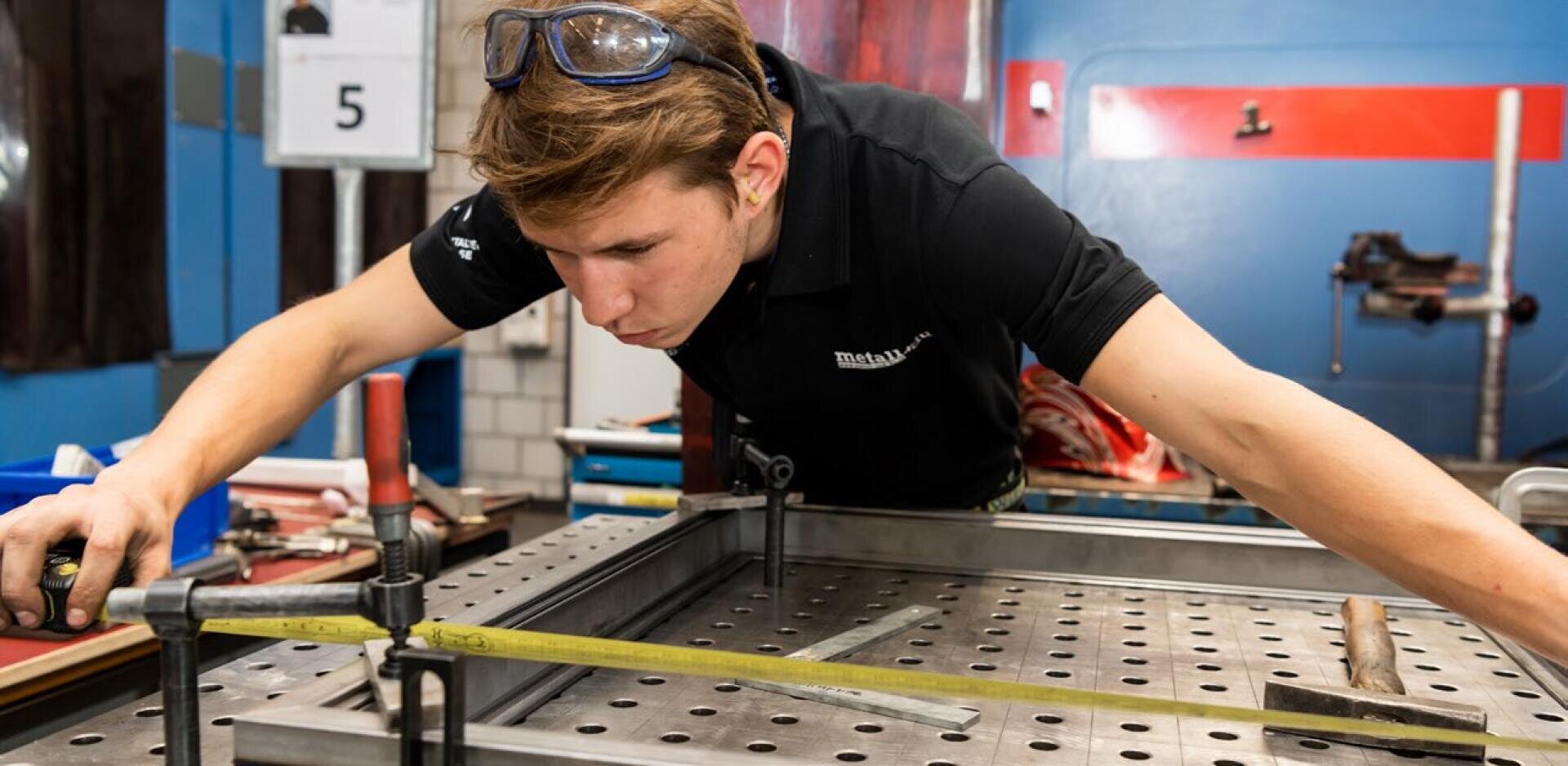 Eine Person, die eine Schutzbrille und ein schwarzes Hemd trägt, misst mit einem Maßband in einer Industriewerkstatt einen Metallrahmen auf einer gelochten Werkbank aus.