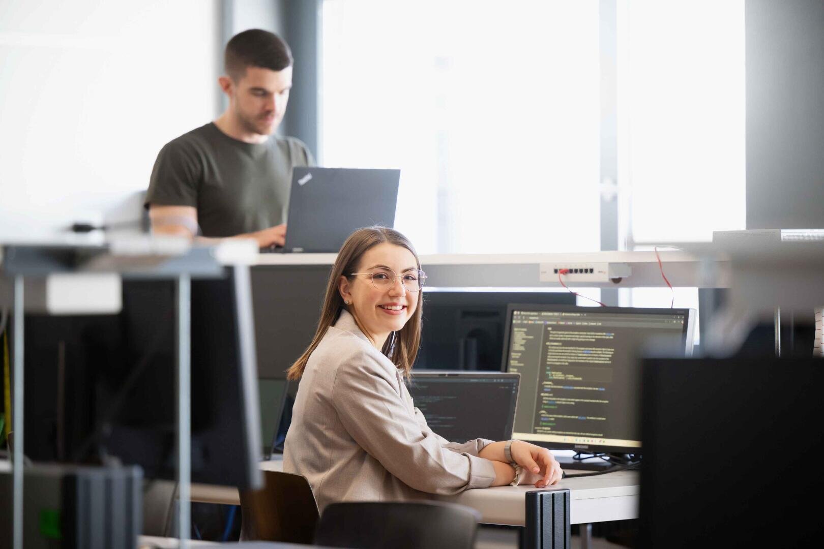 Eine Frau mit Brille sitzt an einem Schreibtisch mit Computermonitoren, die Codes anzeigen, und lächelt in die Kamera. Im Hintergrund arbeitet ein Mann an einem Laptop an einem Stehpult in einem hellen, modernen Büroraum.