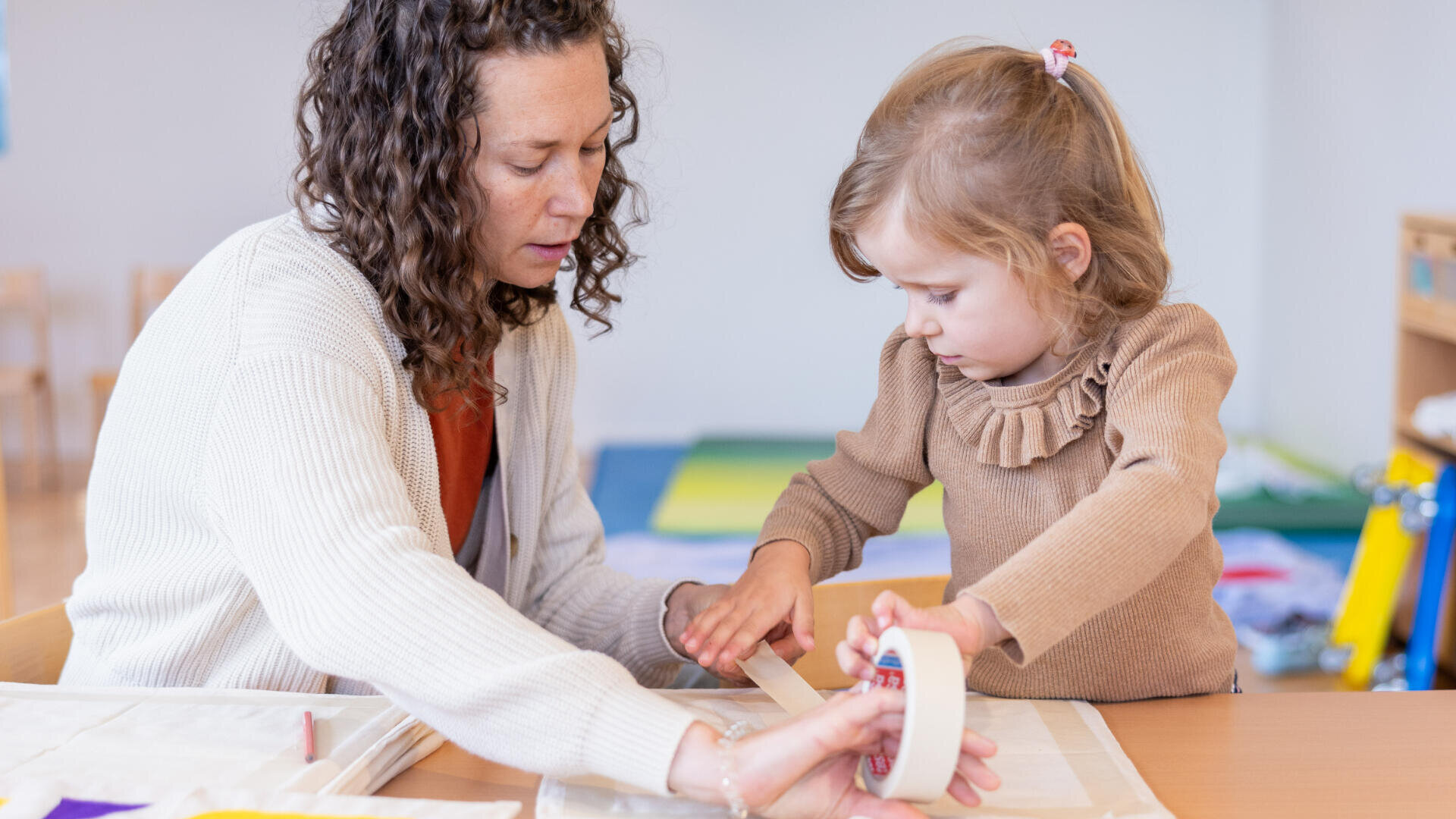 Ein Erwachsener hilft einem jungen Mädchen, Klebeband für ein Bastelprojekt an einem Tisch zu verwenden. Das Kind konzentriert sich auf das Abziehen des Klebebands, während der Erwachsene ihre Hände führt. Im Hintergrund ist ein Klassenzimmer zu sehen.