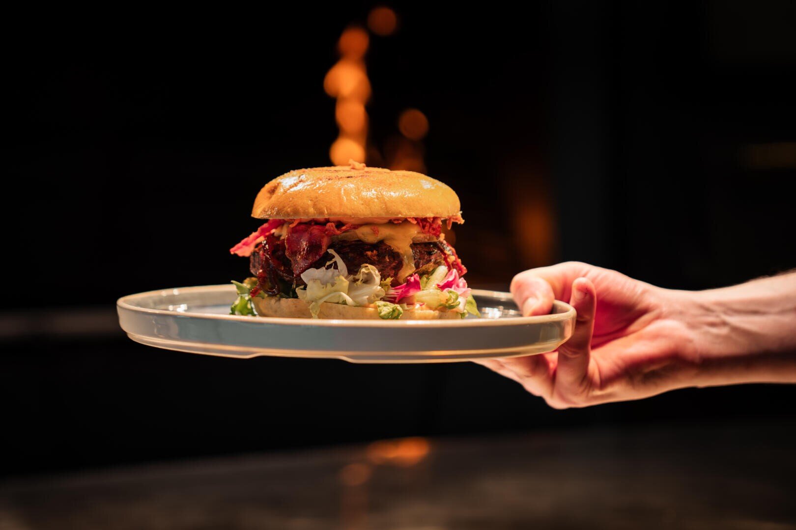 Eine Hand hält einen Teller mit einem Gourmet-Burger, der mit geschmolzenem Käse, Blattgemüse und eingelegtem Gemüse belegt ist, vor einem dunklen Hintergrund, in dem in der Ferne Flammen zu sehen sind.