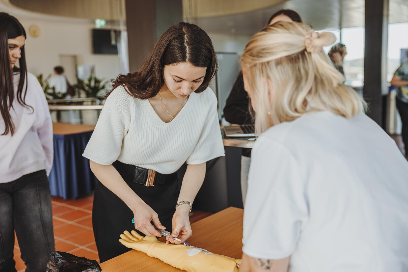 Eine Frau übt das Einführen einer Nadel in den Arm einer medizinischen Übungspuppe, während eine andere Frau aufmerksam zusieht. Im Hintergrund sind in einem hell erleuchteten Raum weitere Personen zu sehen.