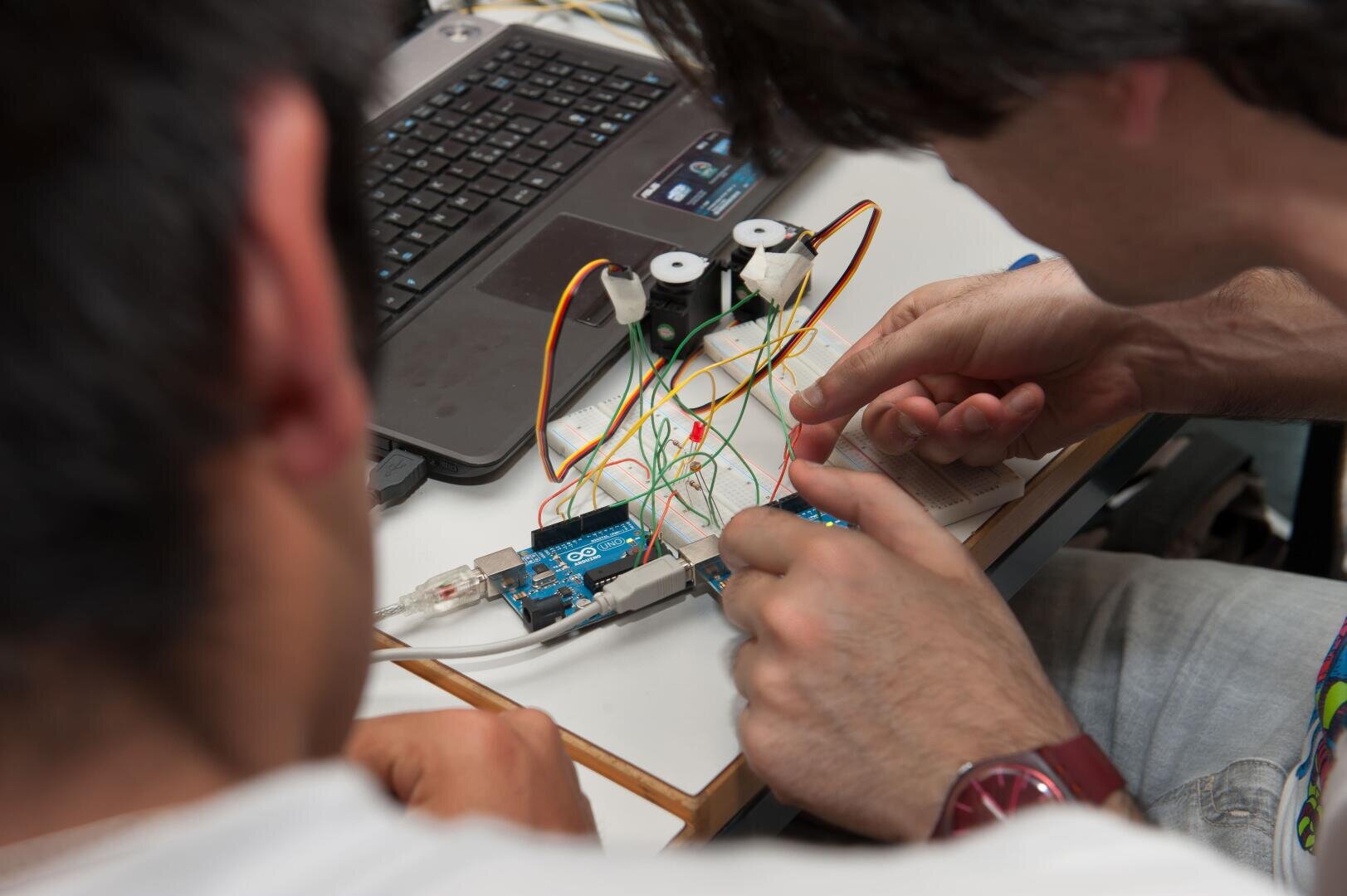Zwei Personen arbeiten an einem kleinen Elektronikprojekt mit bunten Kabeln, einem Arduino-Board und einem Laptop auf einem weißen Schreibtisch. Während sie die Schaltung zusammenbauen, richten sie mit ihren Händen Bauteile aus und schließen Kabel an.
