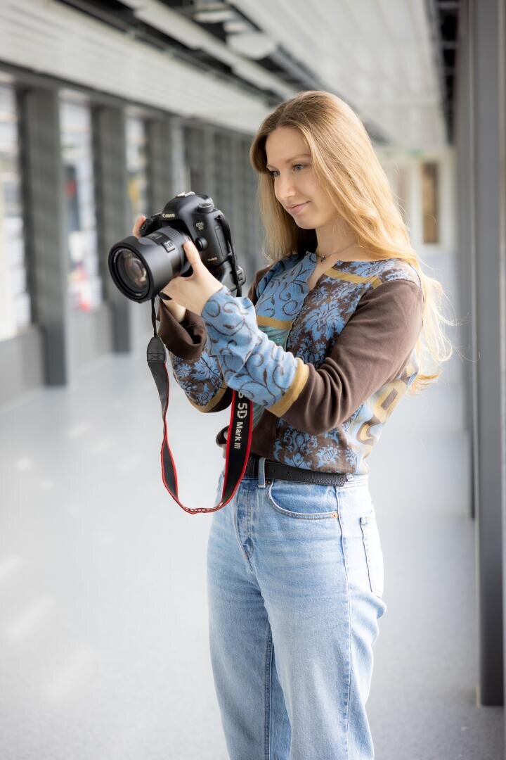 Eine Frau mit langen blonden Haaren, die ein gemustertes langärmeliges Hemd und Jeans trägt, steht in einem hellen Flur und schaut auf den Bildschirm einer DSLR-Kamera, die sie in der Hand hält.