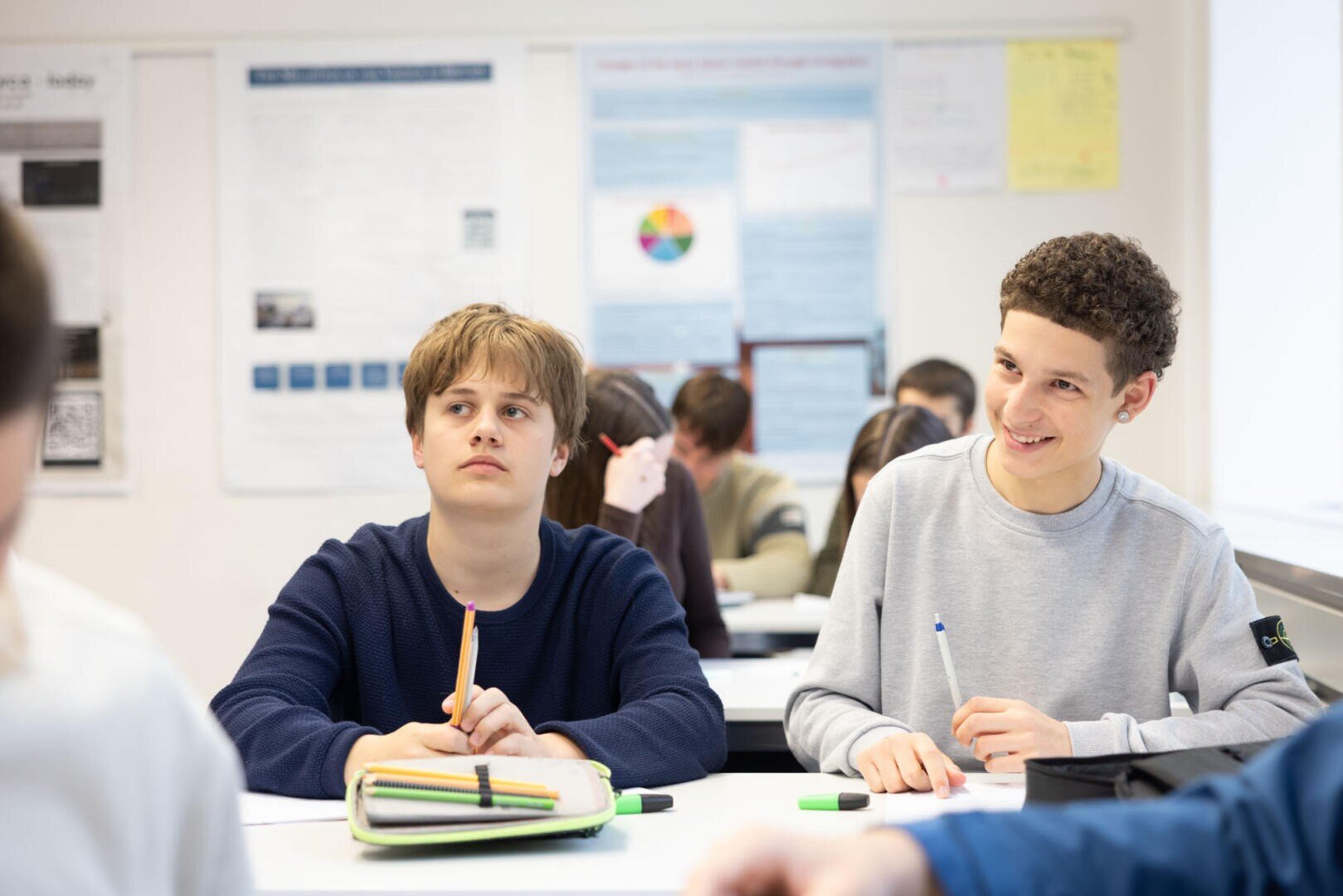Zwei Jungen im Teenageralter sitzen an einem Klassenzimmerpult, der eine schaut aufmerksam, der andere lächelt. Sie haben Hefte, Stifte und Mäppchen vor sich liegen. Im Hintergrund sind andere Schüler und Plakate zu sehen.
