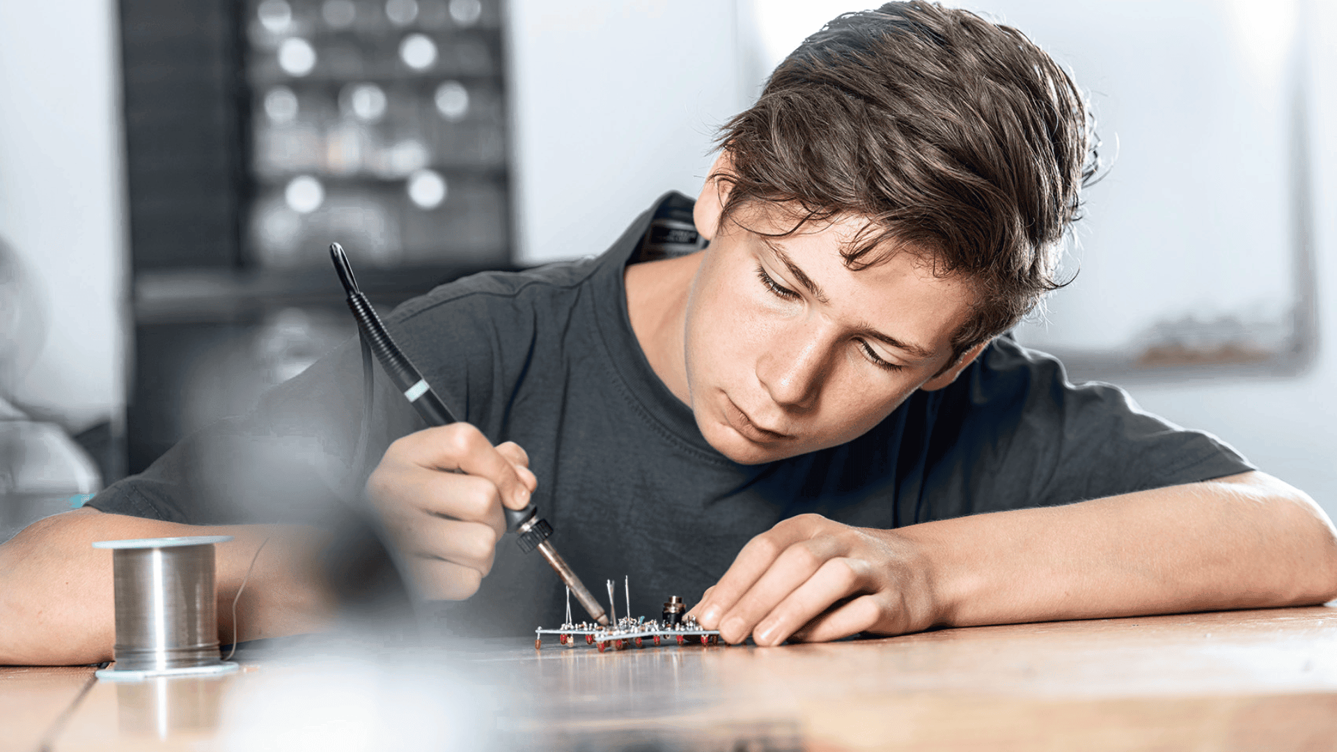 Ein junger Mensch lötet an einem Schreibtisch sorgfältig elektronische Bauteile auf eine Leiterplatte, eine Spule mit Lötdraht liegt in der Nähe.
