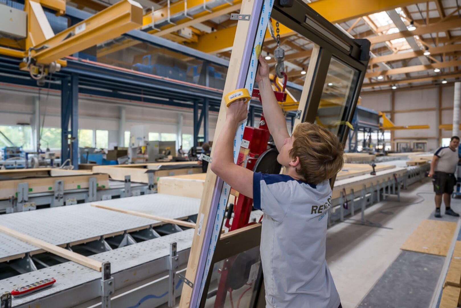 Eine Person in einer Fabrik verwendet ein Werkzeug, um einen großen Fensterrahmen auf einer Fertigungsstraße zu messen oder einzustellen. Die industrielle Umgebung besteht aus großen Maschinen, Werkbänken und hohen Decken mit heller Beleuchtung.