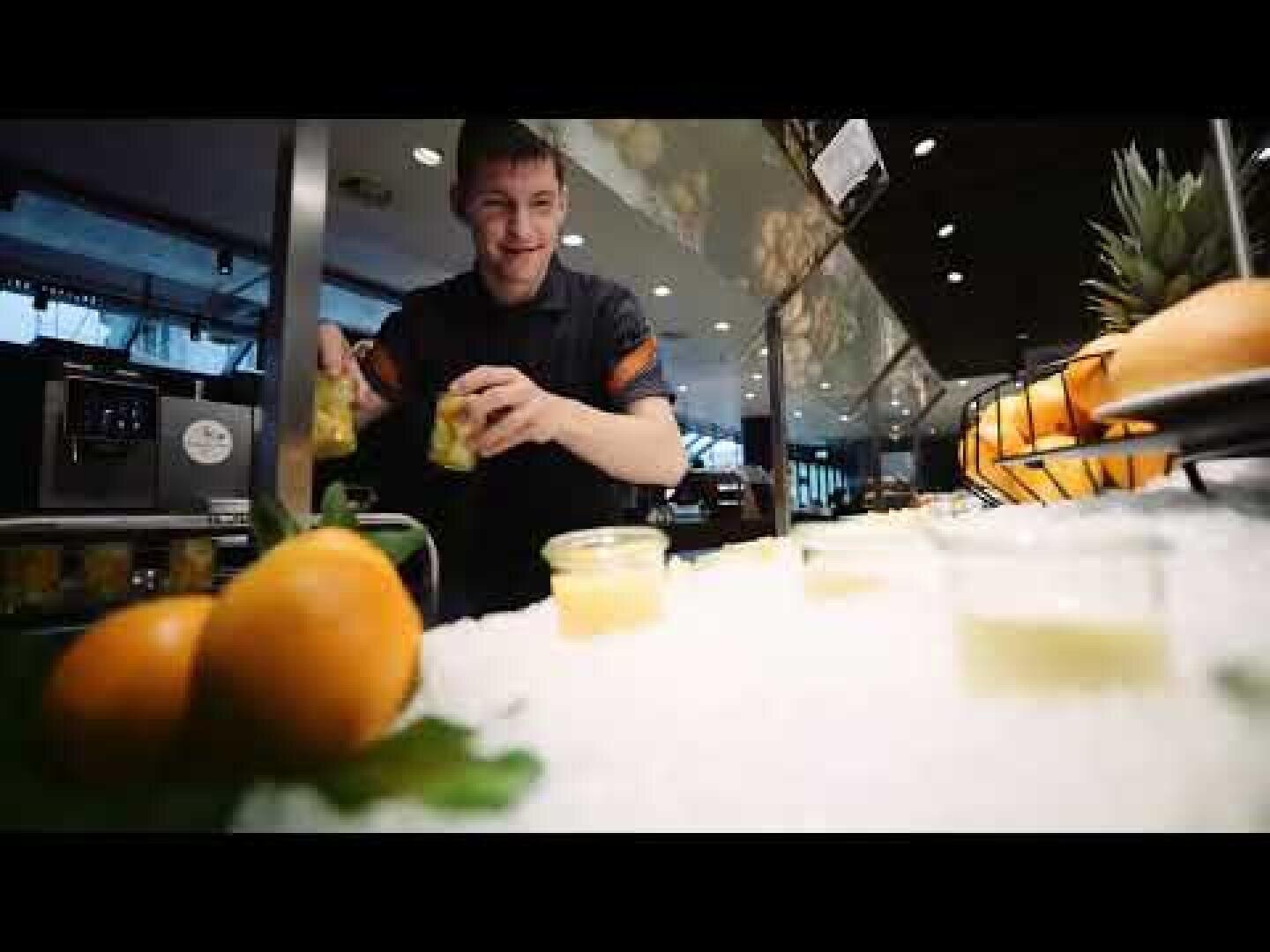 Eine Person hinter einer Theke stellt Schälchen mit Obstsalat auf, daneben liegen Orangen und Ananas. Die Szene scheint in einer modernen Cafeteria oder einem Buffet zu spielen.