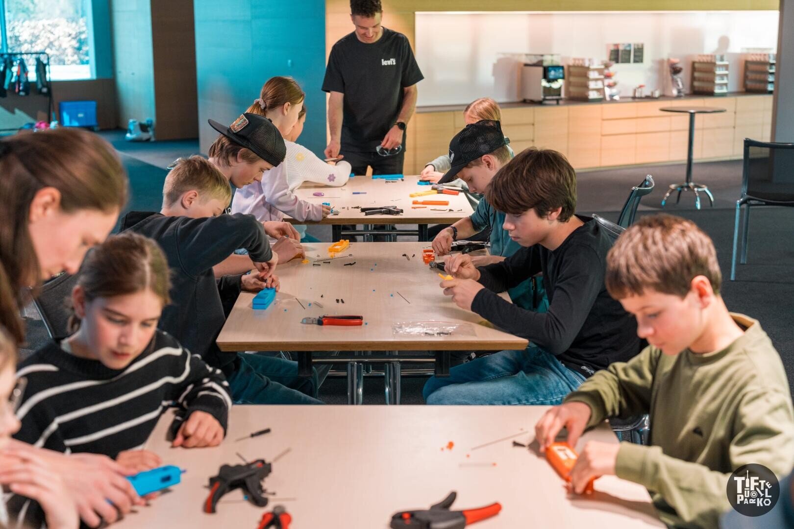 Kinder und Erwachsene sitzen um einen Tisch herum und basteln mit Heißklebepistolen und kleinen Werkzeugen in einem hell beleuchteten Klassenzimmer oder Werkraum.