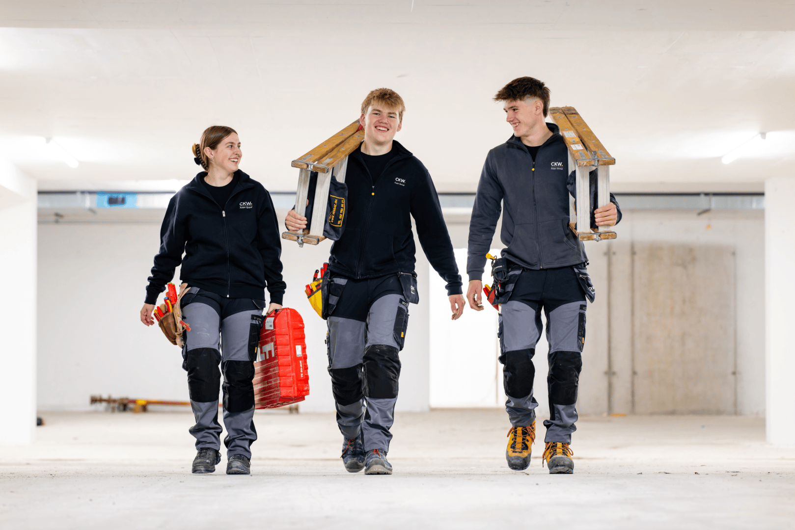 Drei Bauarbeiter in einheitlichen Uniformen gehen lächelnd gemeinsam durch einen Innenraum. Zwei tragen Leitern auf den Schultern, während der dritte einen roten Werkzeugkasten trägt. Der Raum wirkt unfertig, mit einem Betonboden und weißen Wänden.