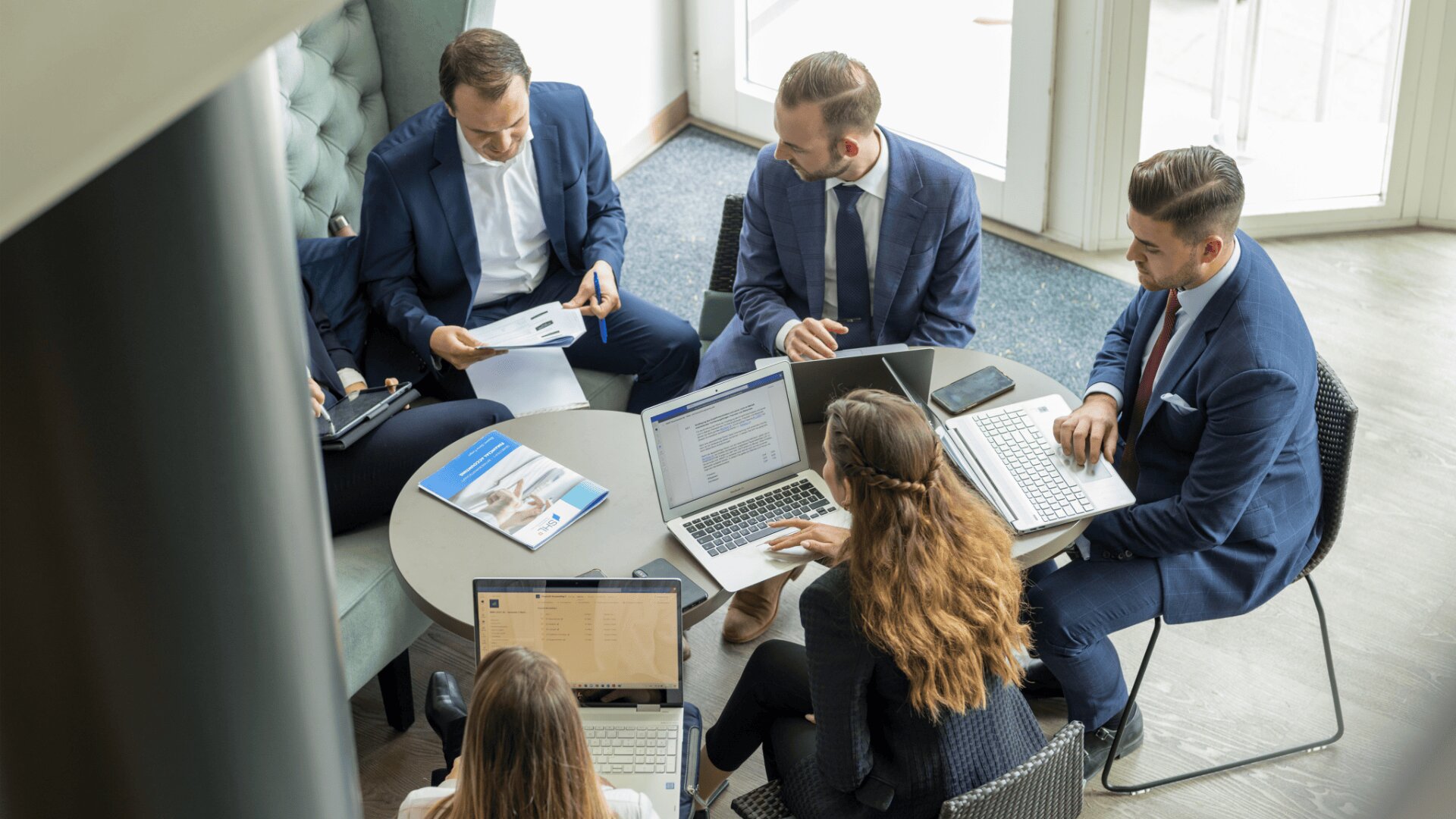 Fünf Personen in Geschäftskleidung sitzen um einen kleinen Tisch mit Laptops, Dokumenten und Tablets und führen eine Besprechung oder ein Gespräch in einer modernen Büroumgebung mit natürlichem Licht.