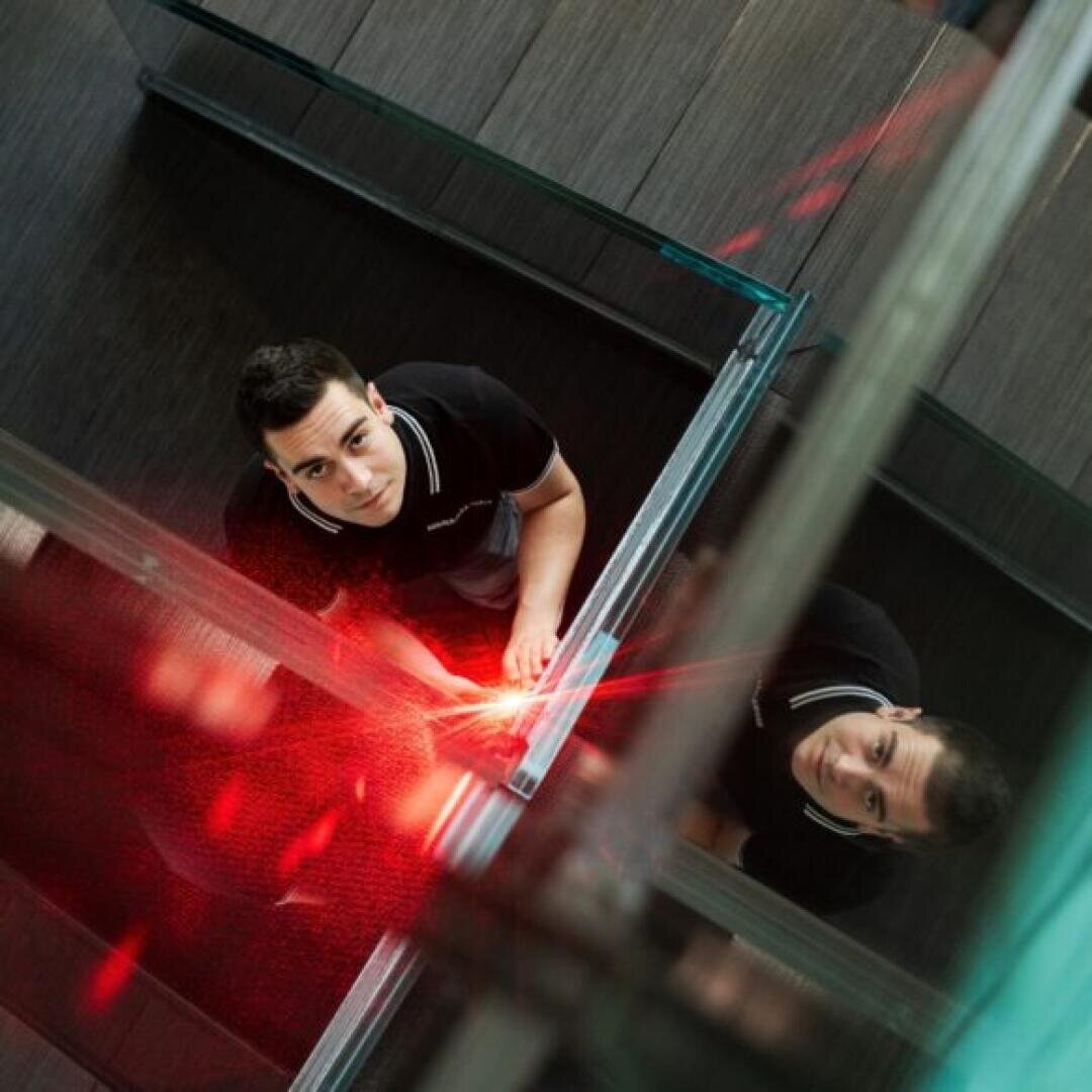 Ein junger Mann in einem schwarzen Hemd steht neben einer Glaswand und schaut nach oben. Sein Spiegelbild ist im Glas sichtbar, und rotes Licht leuchtet in seiner Nähe und schafft eine moderne, dynamische Atmosphäre.