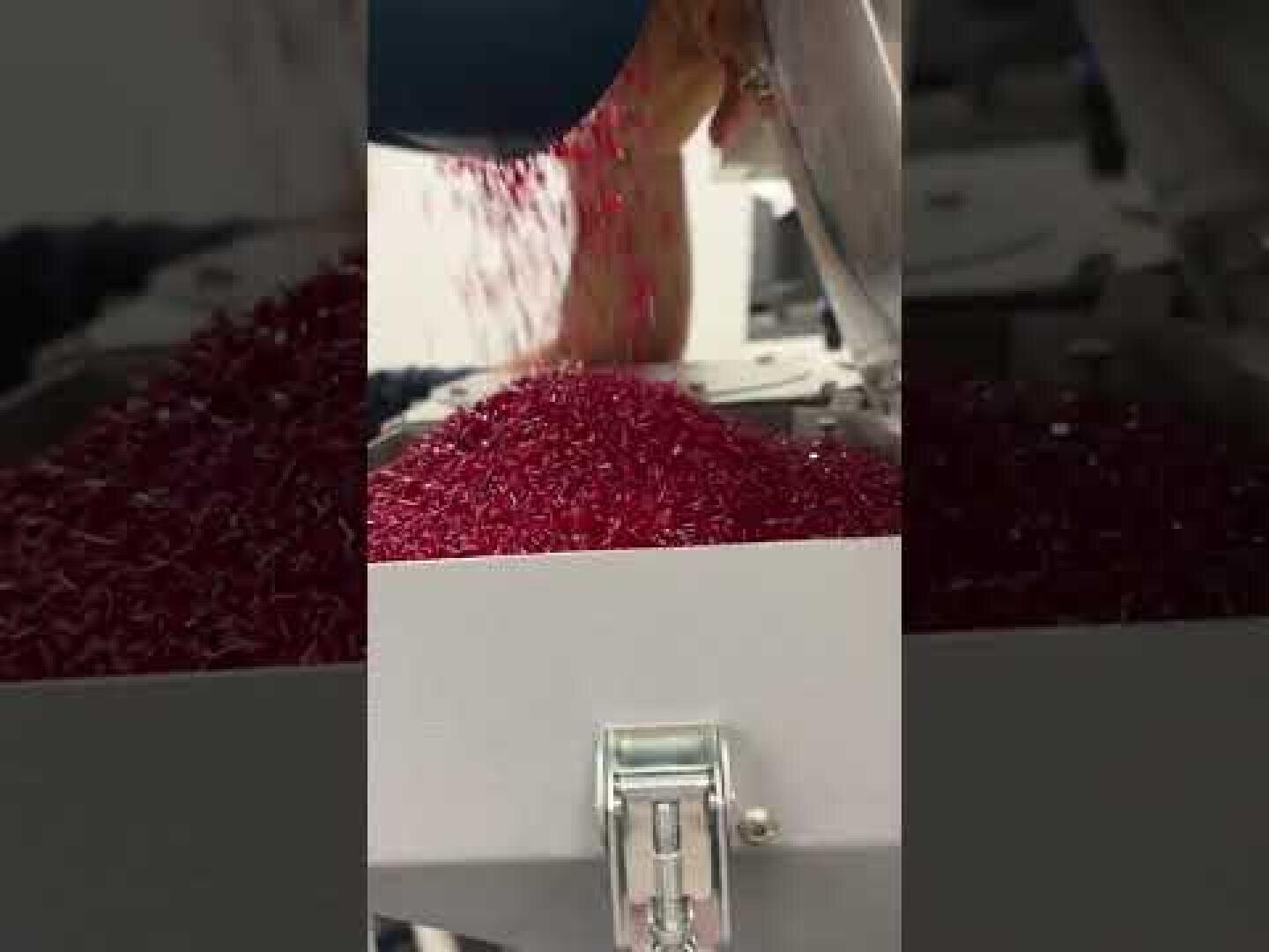 Rote Kunststoffgranulate werden aus einem Behälter in eine Sortier- oder Verarbeitungsmaschine geschüttet, an deren Vorderseite ein Metallverschluss zu sehen ist.