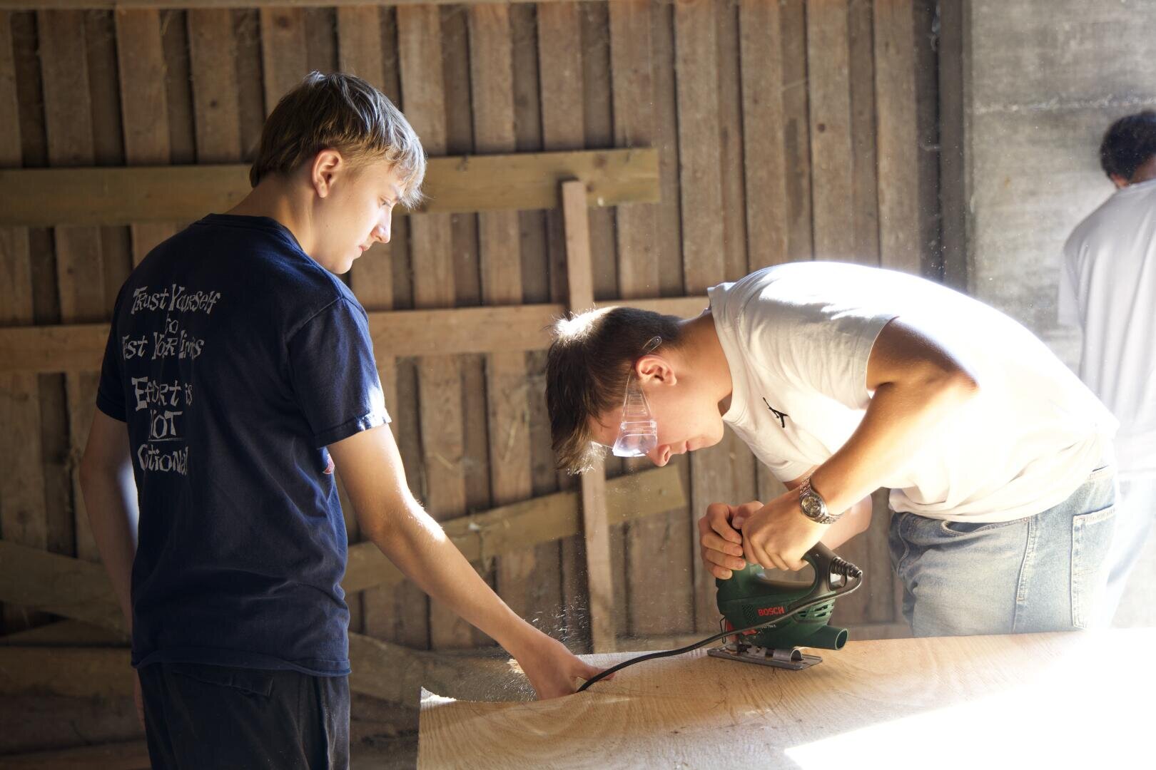 Zwei junge Leute arbeiten an einem Holzbearbeitungsprojekt; einer arbeitet mit einer Motorsäge an einem Holzbrett und trägt eine Schutzbrille, während der andere in der Nähe steht und das Brett in einer sonnendurchfluteten Werkstatt festhält.